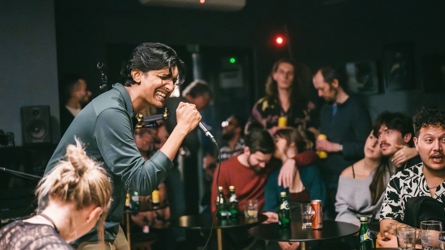 Singer performing with microphone at a lively indoor music event with crowd gathered around tables.