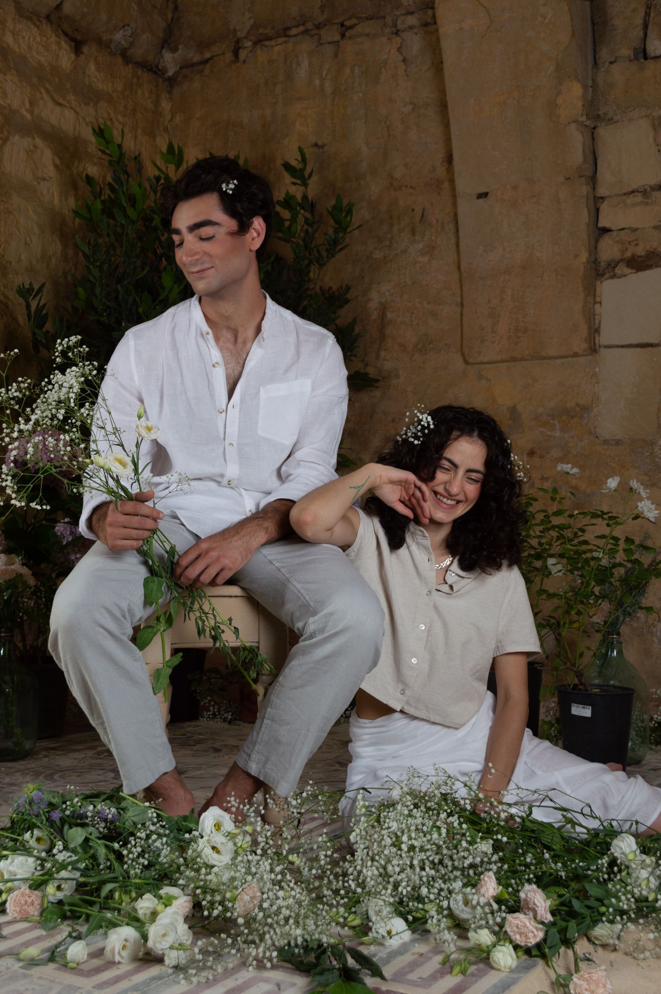 Couple in white linen relaxing among fresh flowers and greenery in a rustic stone interior.