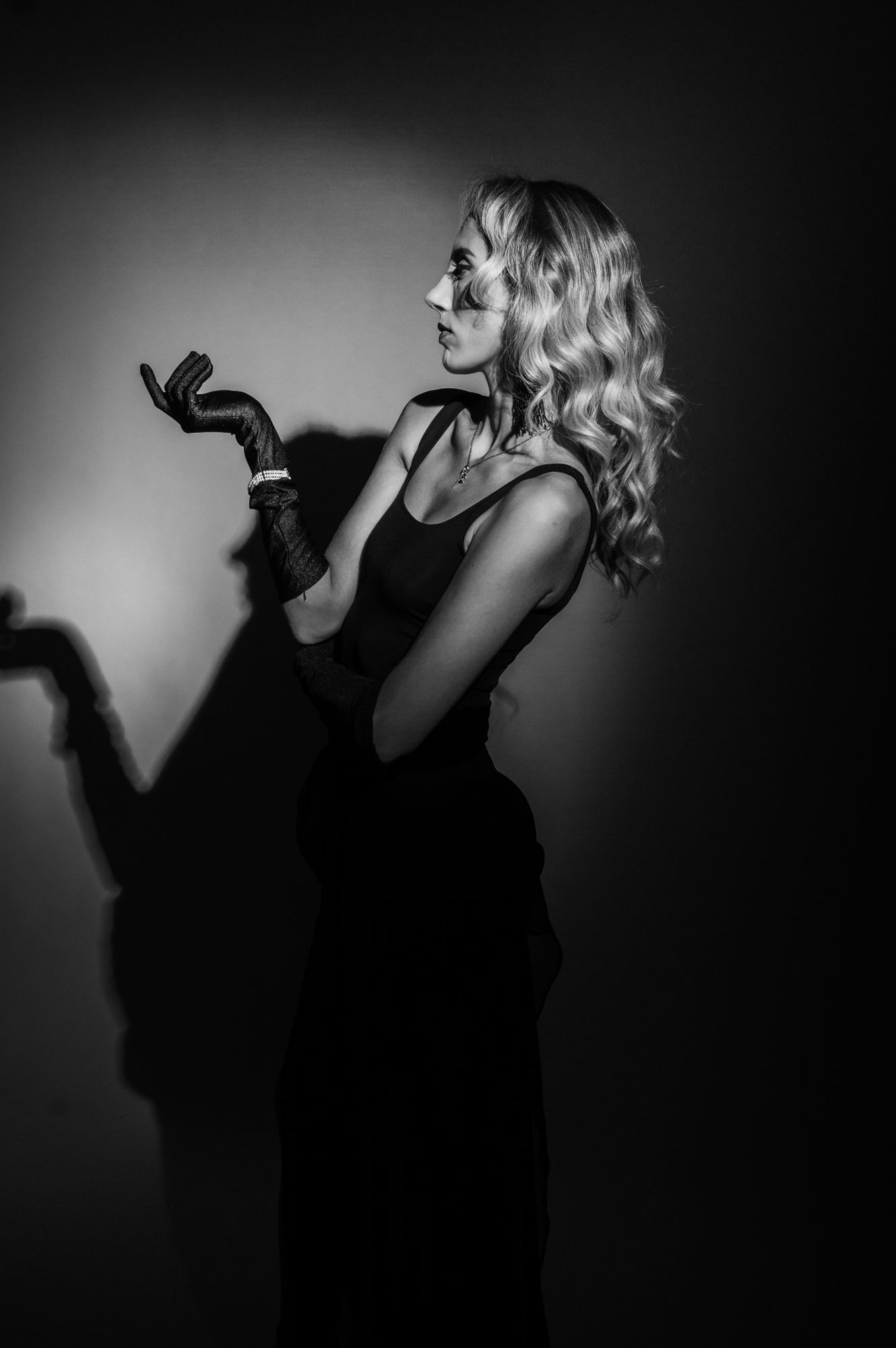 Black and white portrait of a blonde woman in elegant attire with long gloves against dark background.
