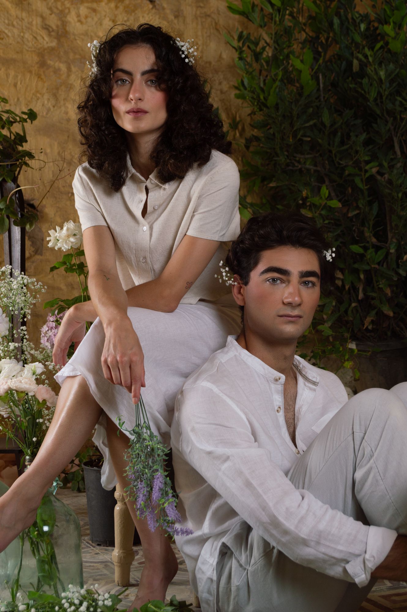 Young couple in white linen clothing posing among wildflowers against a warm stone wall backdrop.