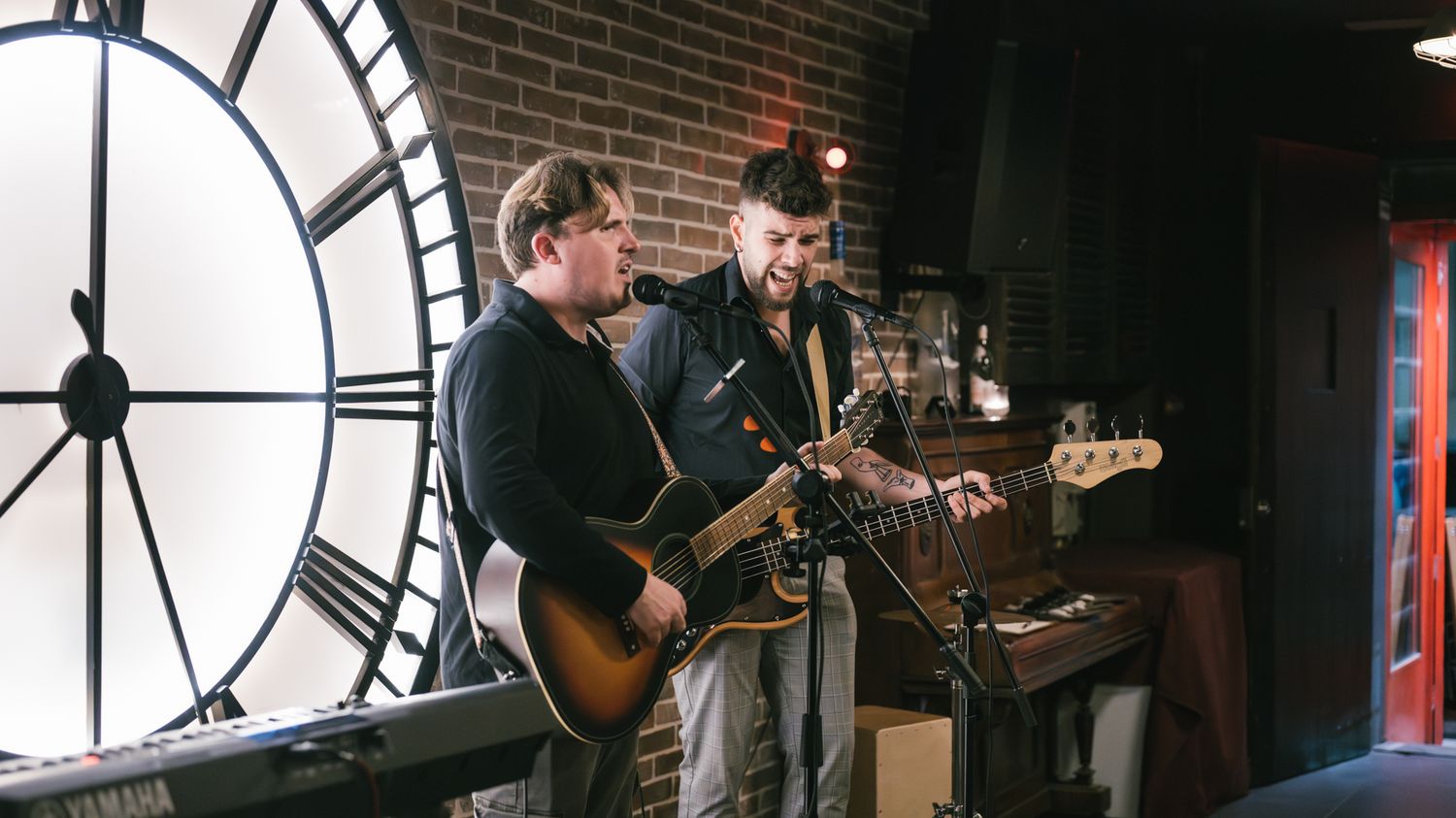 Two musicians playing guitars on stage in front of a large clock on a brick wall.