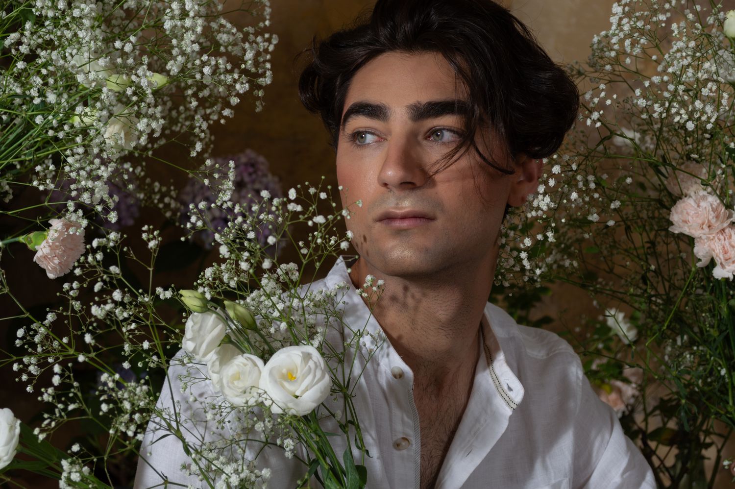 A young man in a white shirt gazes upward surrounded by lush white and pastel floral arrangements.