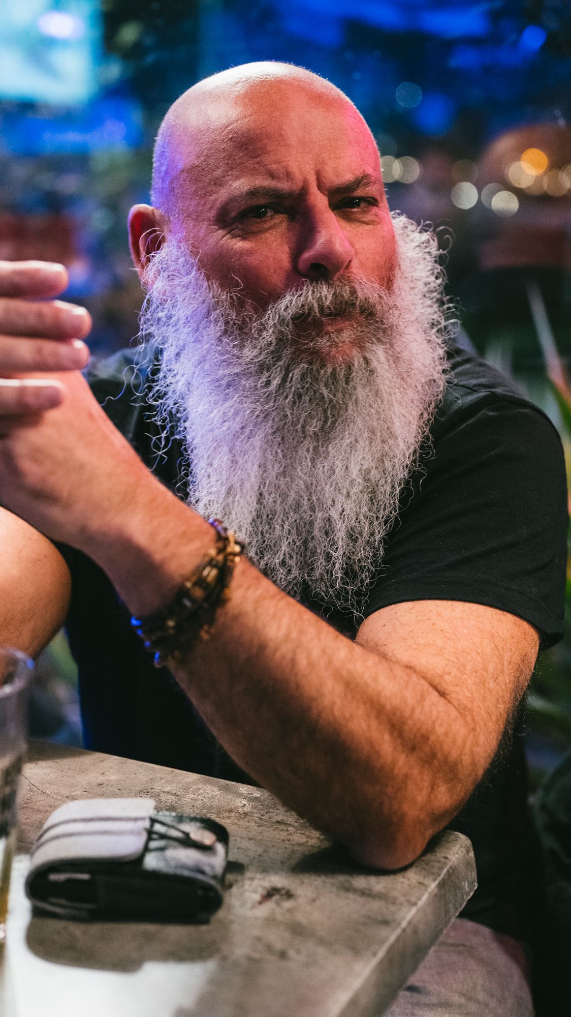 Bald man with long white beard wearing a black t-shirt and beaded bracelet sitting at a bar.