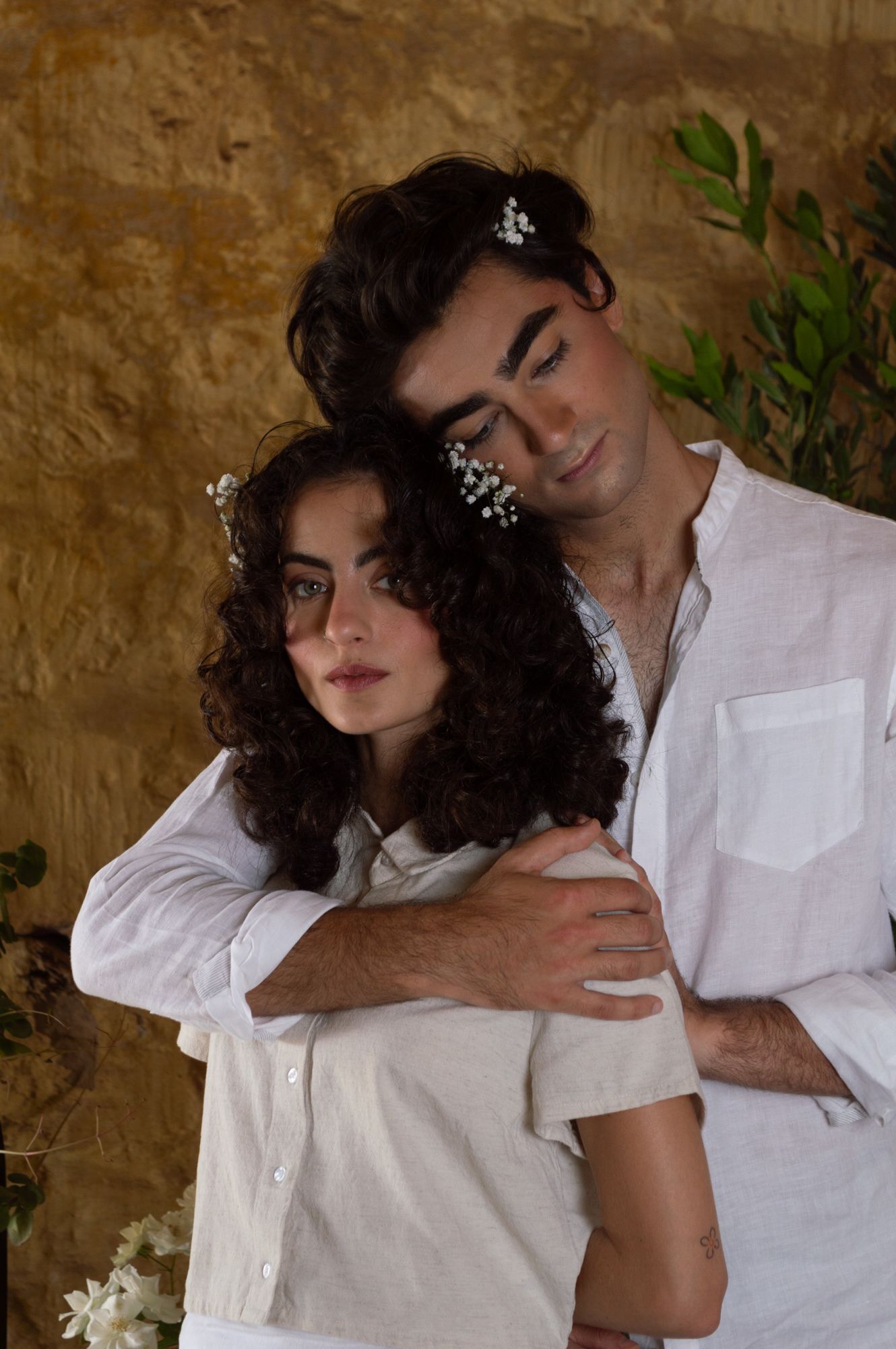Intimate couple portrait in white linen clothing embracing against a rustic golden stone wall.