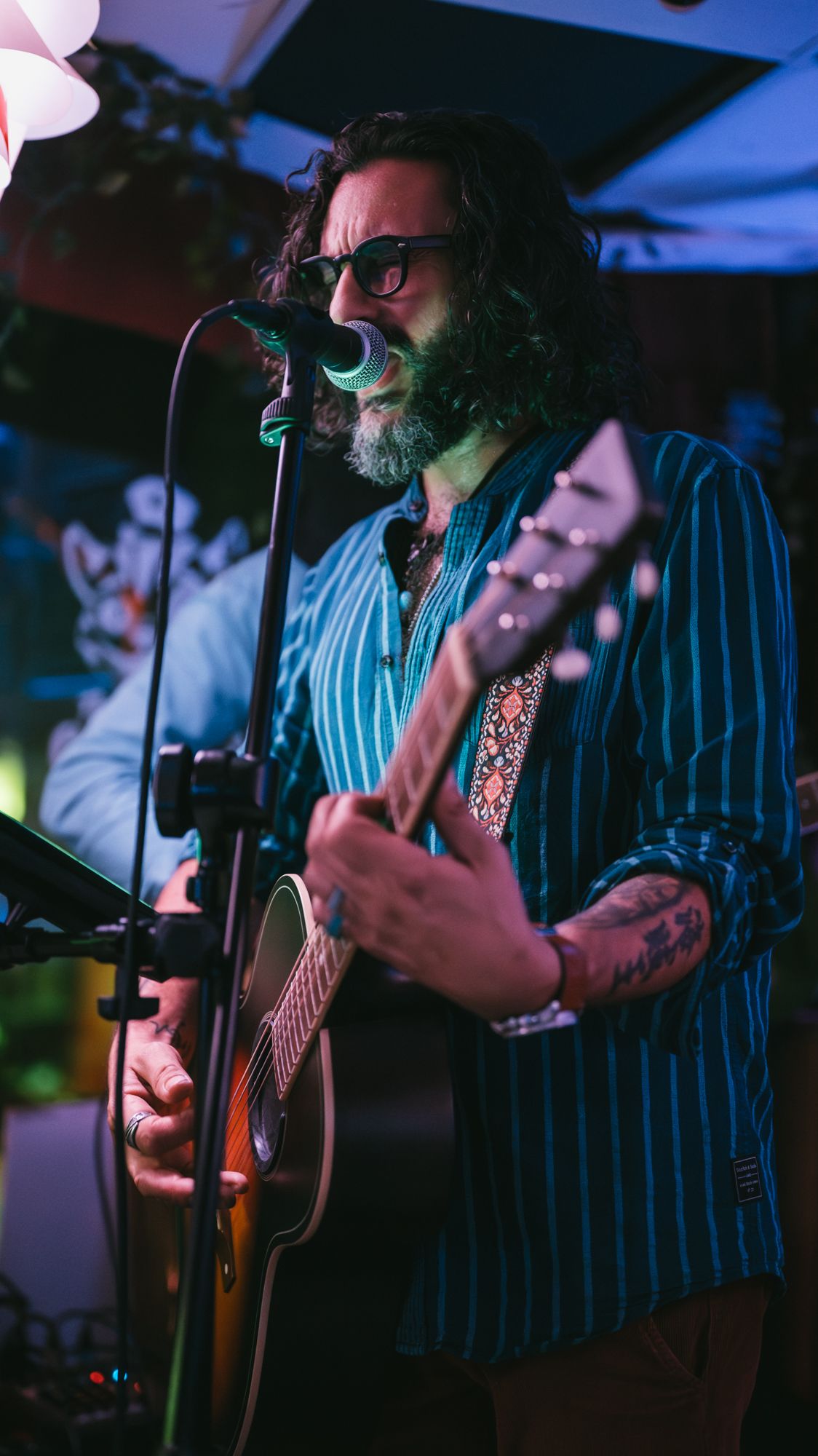 Bearded tattooed man with glasses singing and playing acoustic guitar on stage.