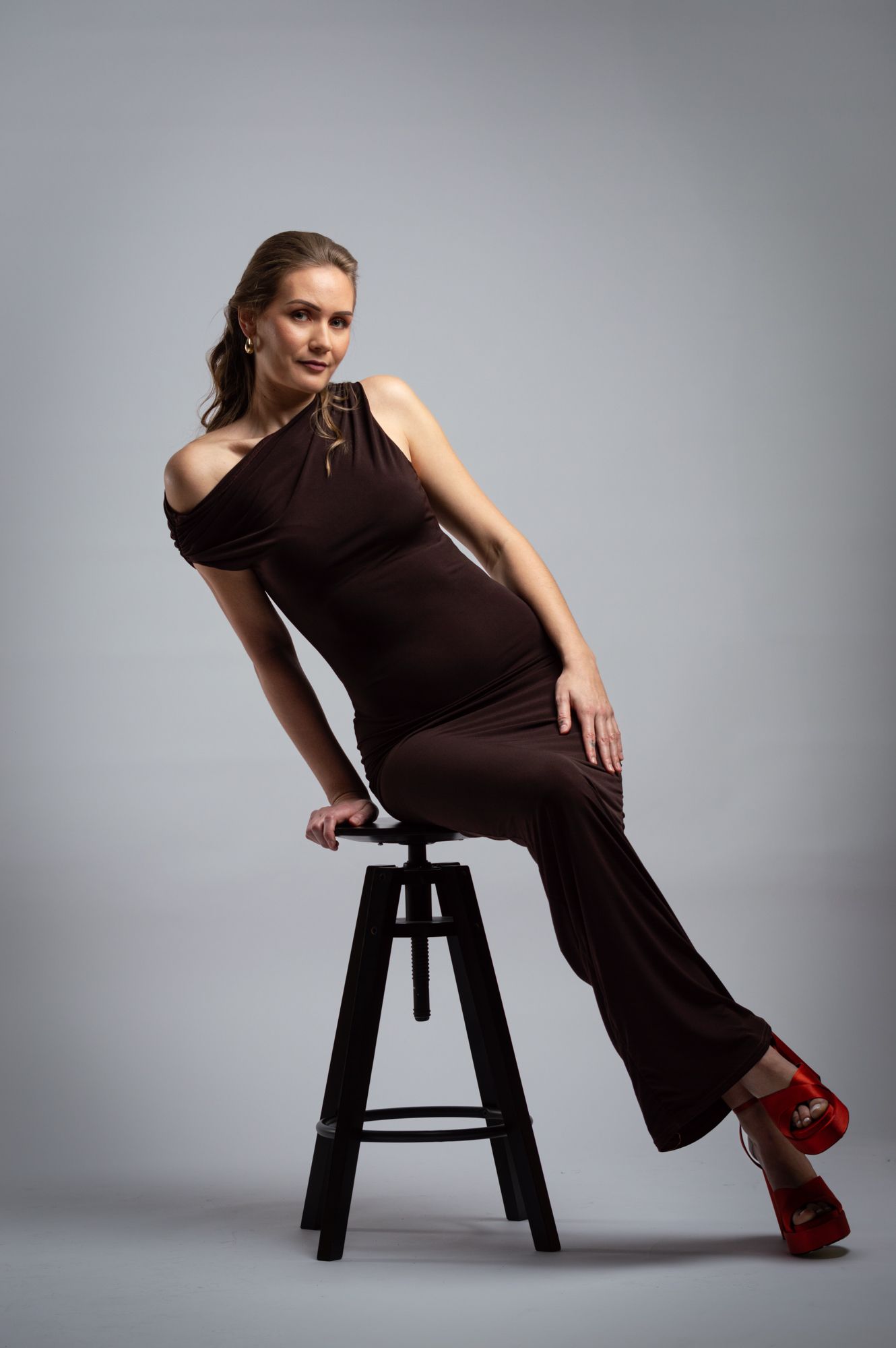 Woman in a brown halter jumpsuit and red heels posing on a stool against a gray background.