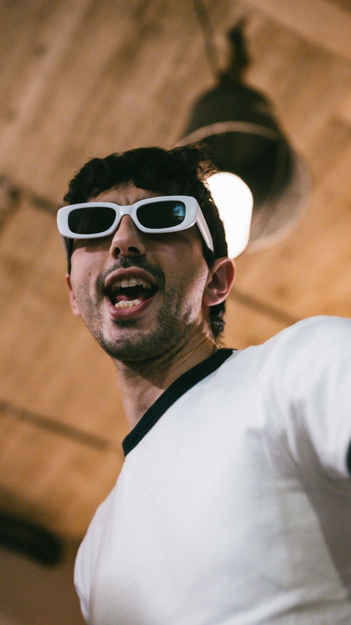 Young man wearing white rectangular sunglasses takes a low-angle selfie under a warm ceiling light.