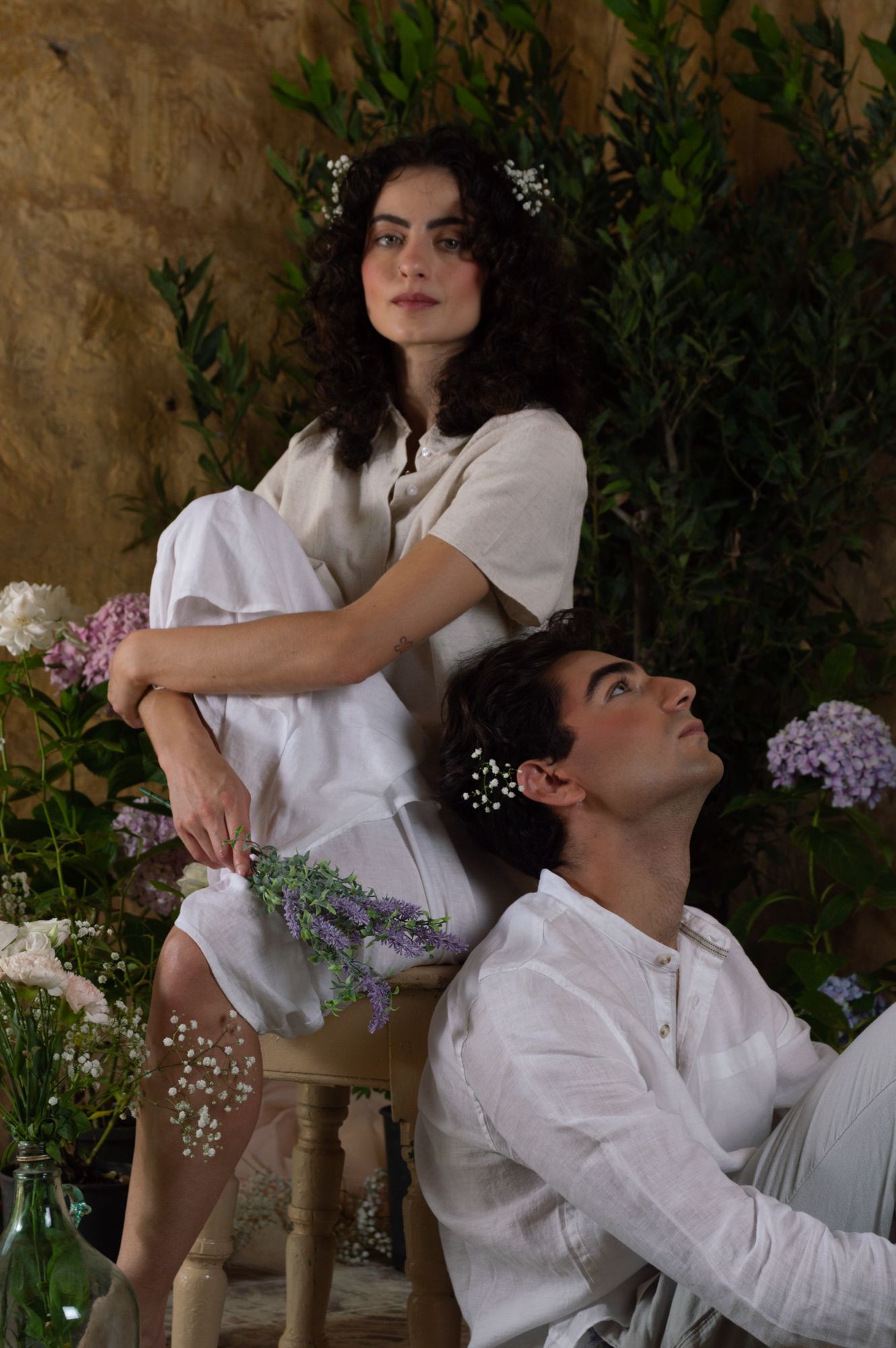 A young woman sits elevated on a chair while a man sits below her among flowers in a rustic floral setting.