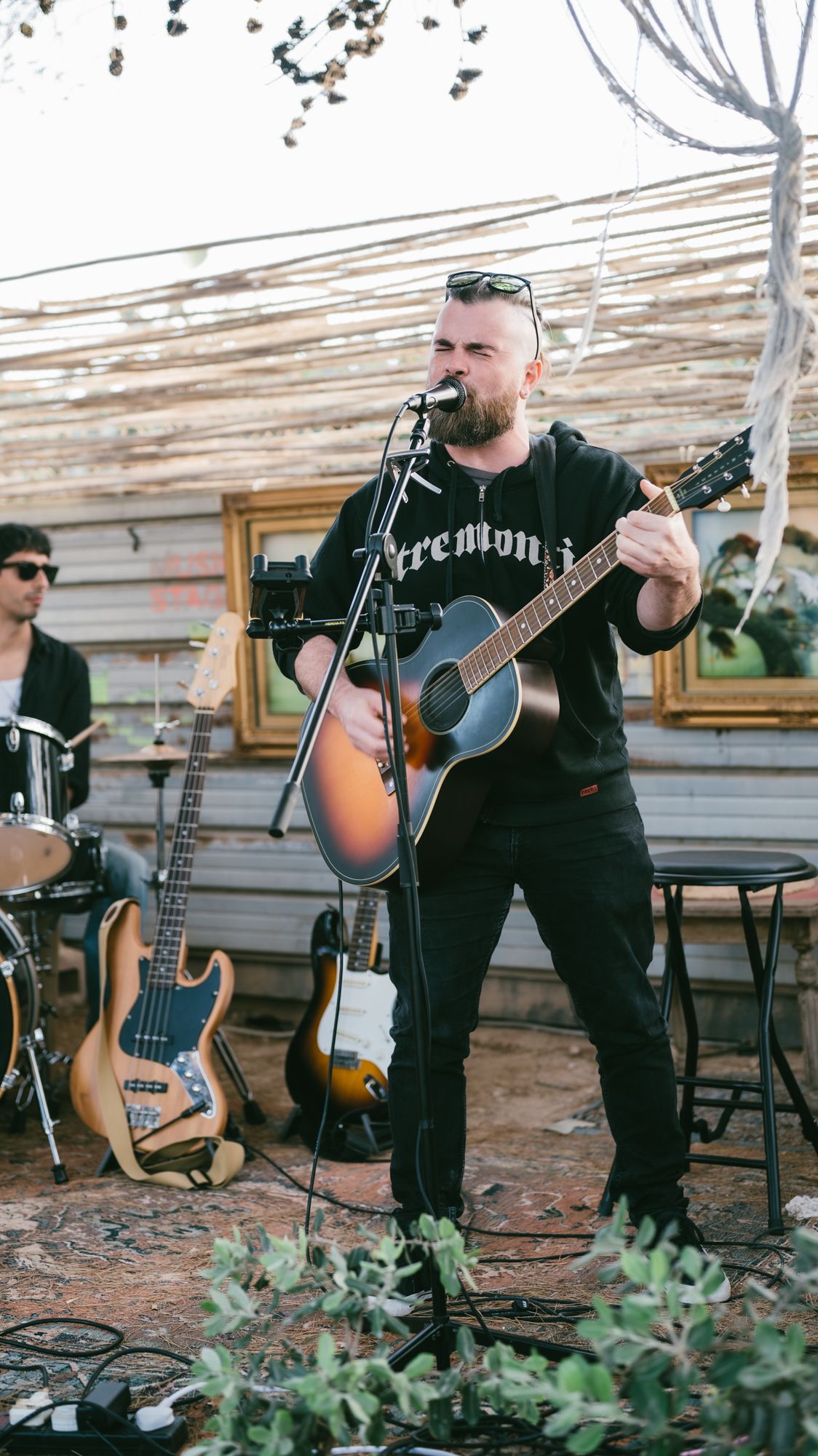Bearded man in black hoodie playing acoustic guitar and singing into microphone outdoors at a rustic venue.