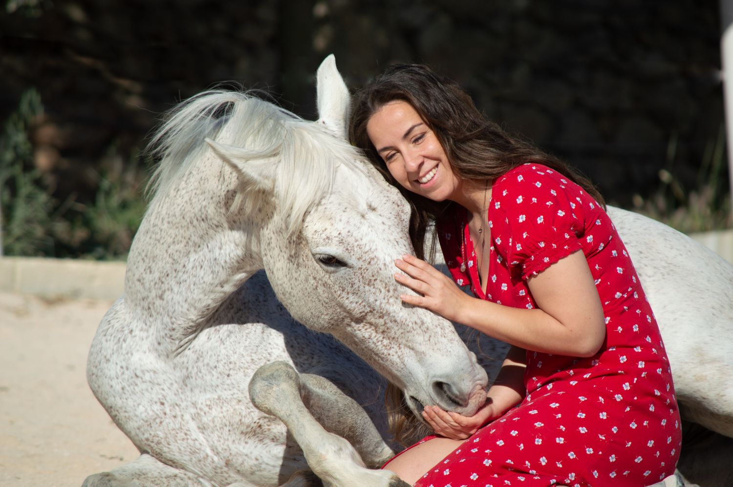 Woman in red dress smiling while hugging a white horse sitting on sandy ground outdoors.