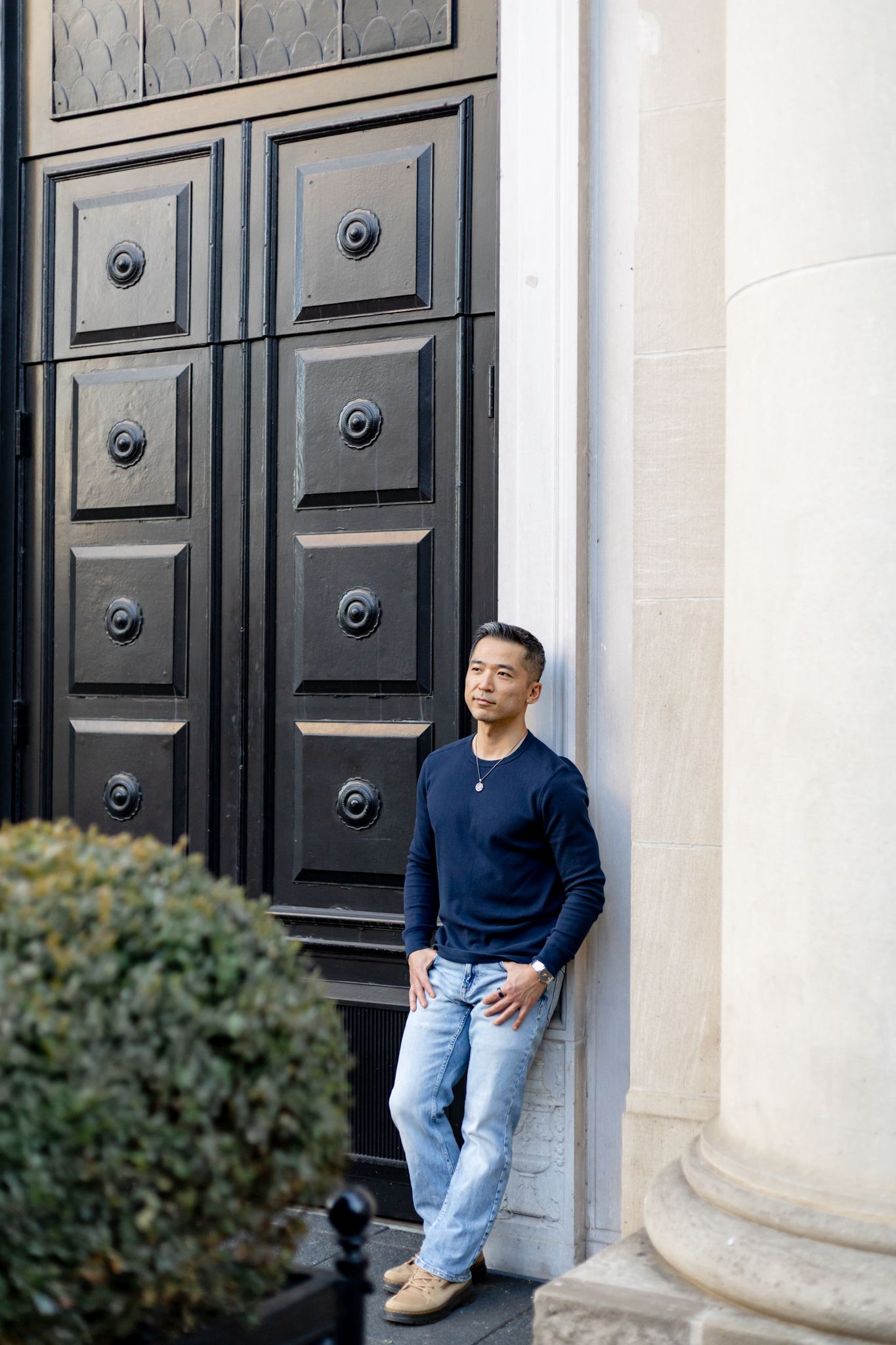 Person in navy sweater and light jeans sits casually against white stone column near black doors.