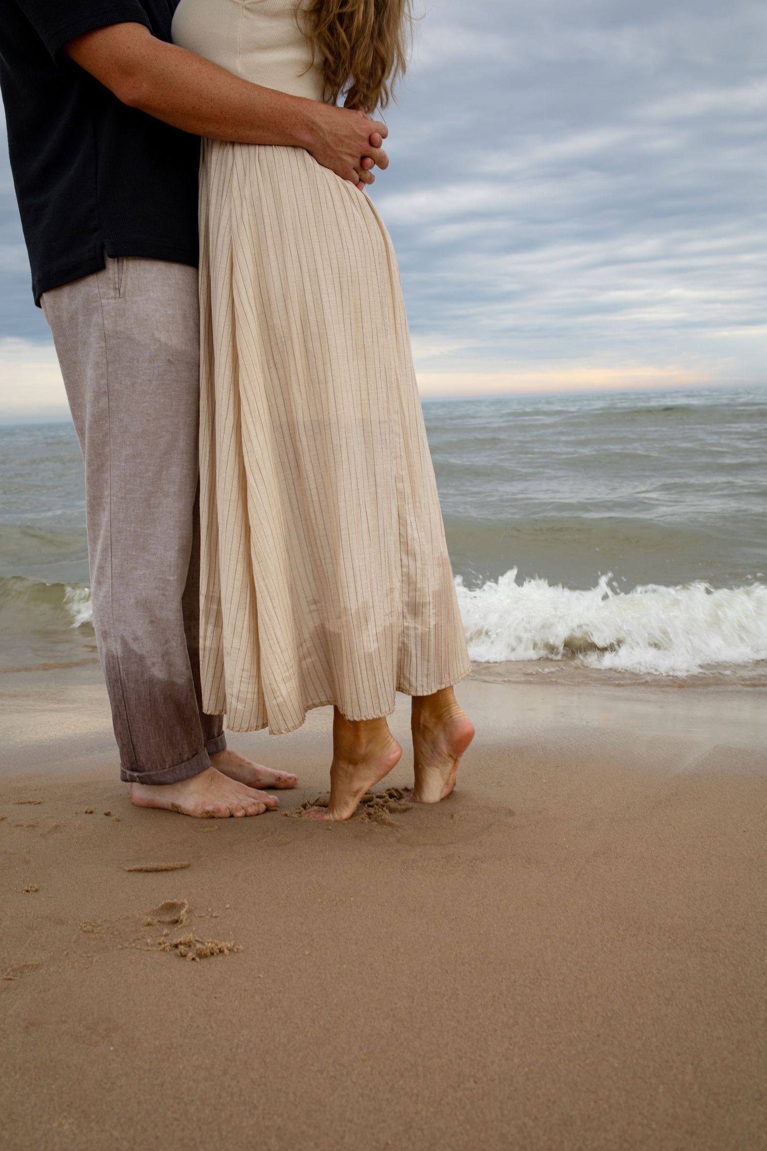 Bare feet standing close together on a sandy beach with gentle waves in the background.