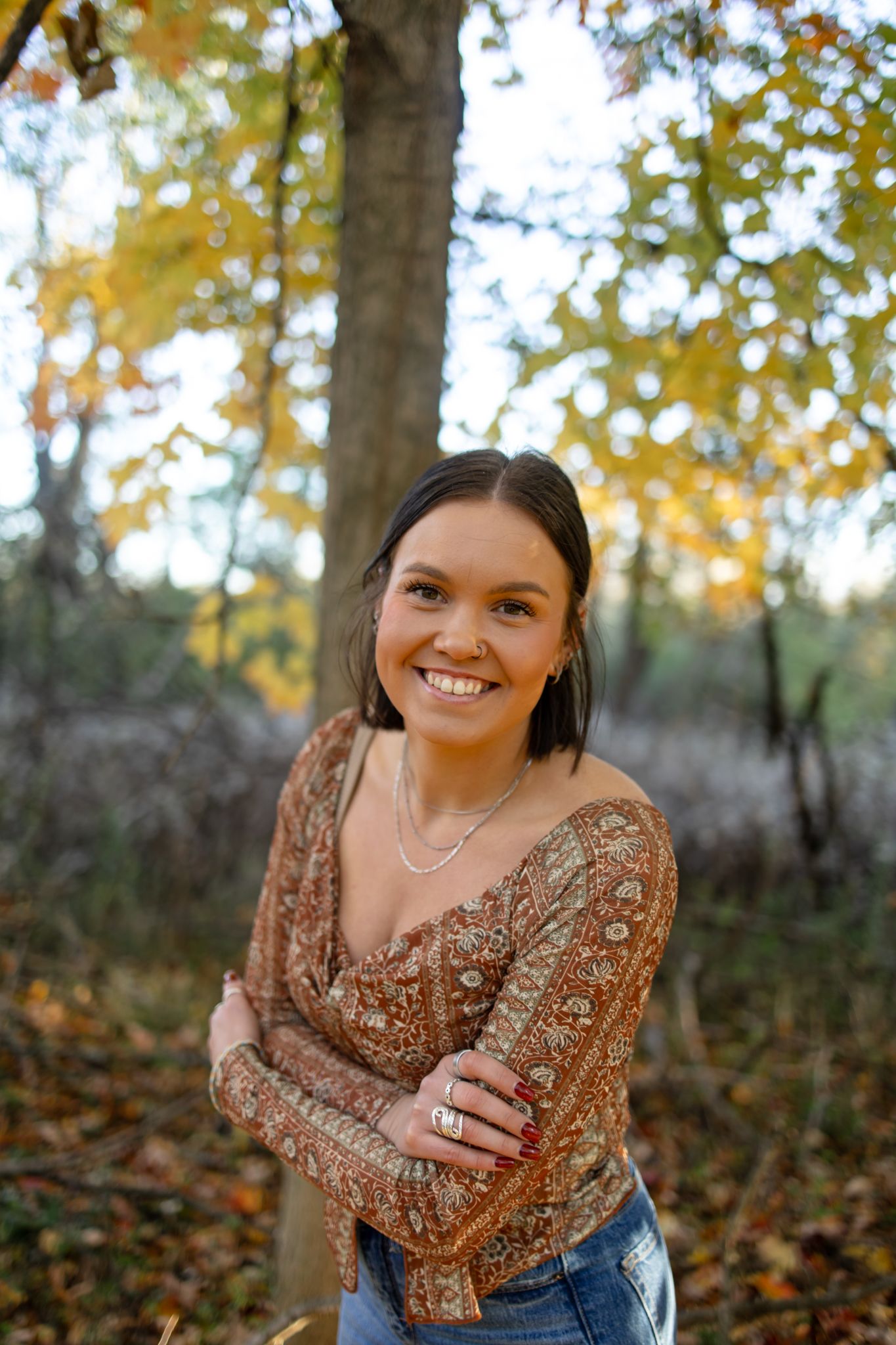 Person in brown patterned top posing outdoors with autumn foliage in background.