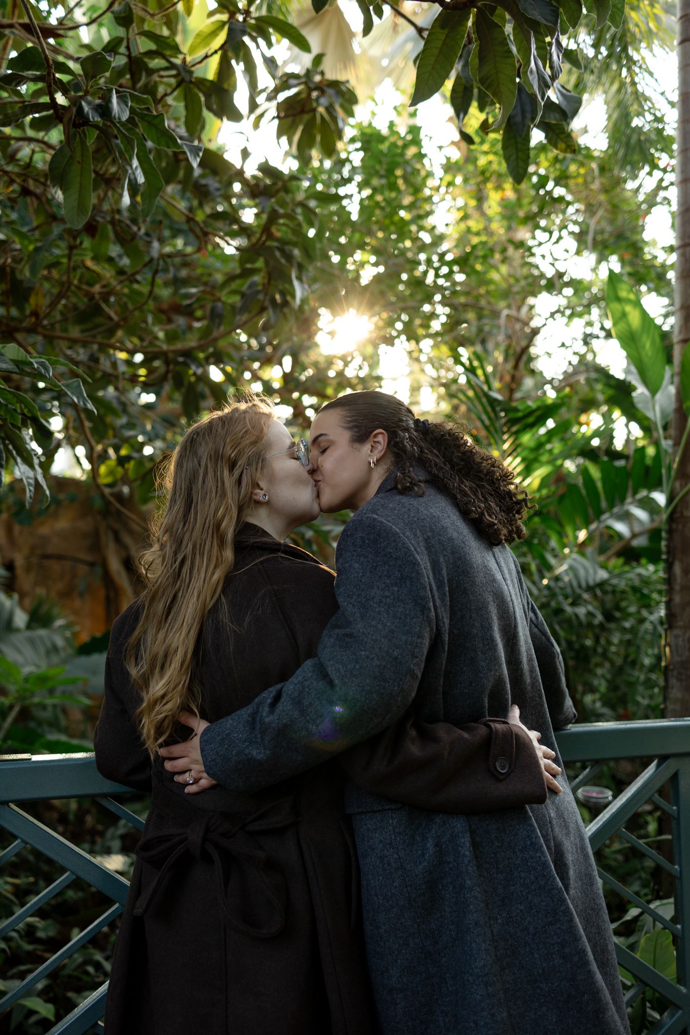 A couple shares a romantic kiss in a lush garden setting with sunlight filtering through green foliage.