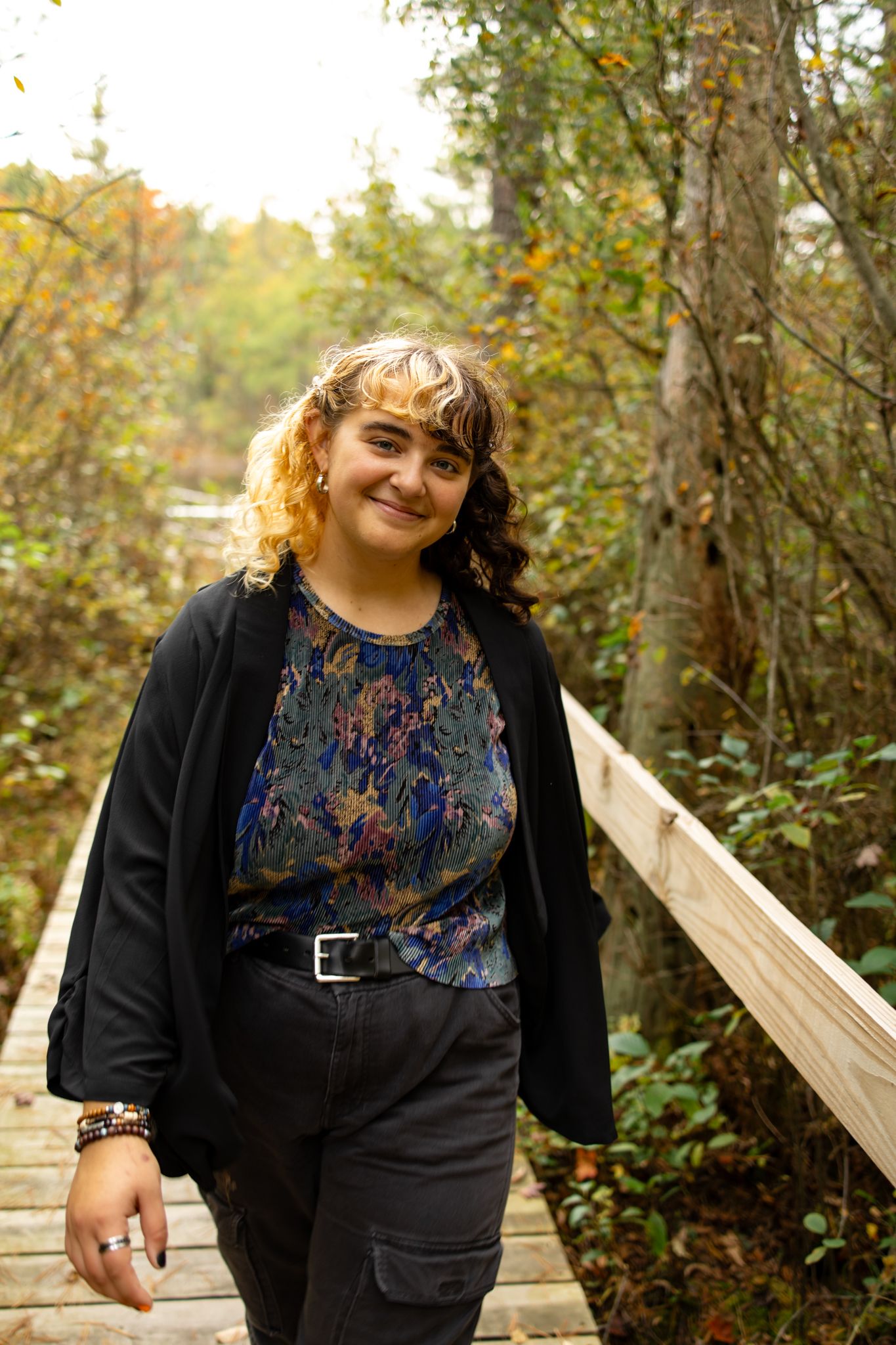 Person in black jacket and patterned top walking along wooden boardwalk surrounded by autumn foliage.