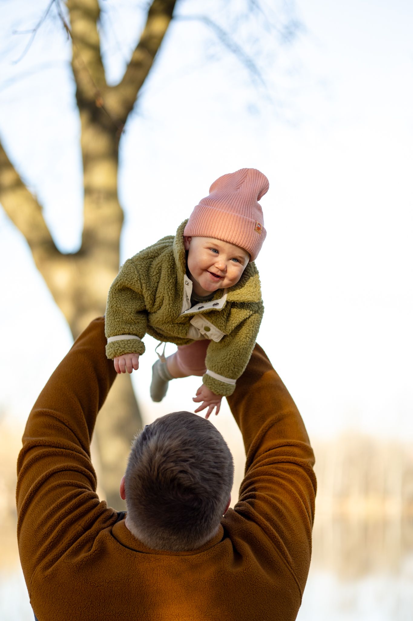 A playful moment lifting a baby wearing a pink beanie and olive green jacket against a bright sky.