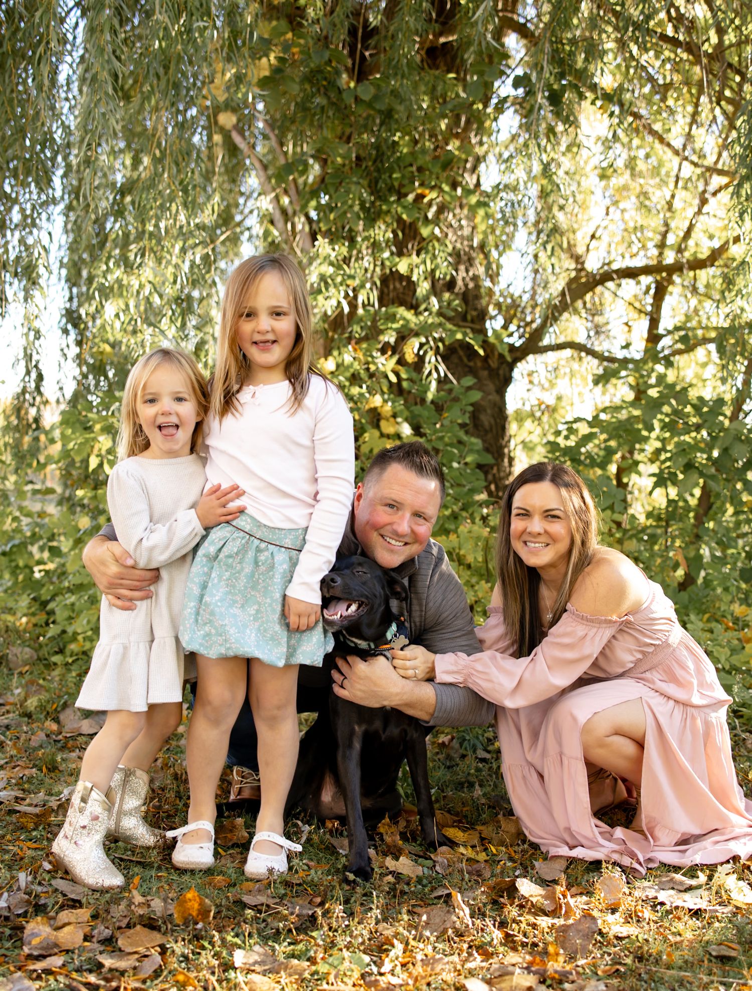 Family with children and dog pose together under willow trees in autumn setting.