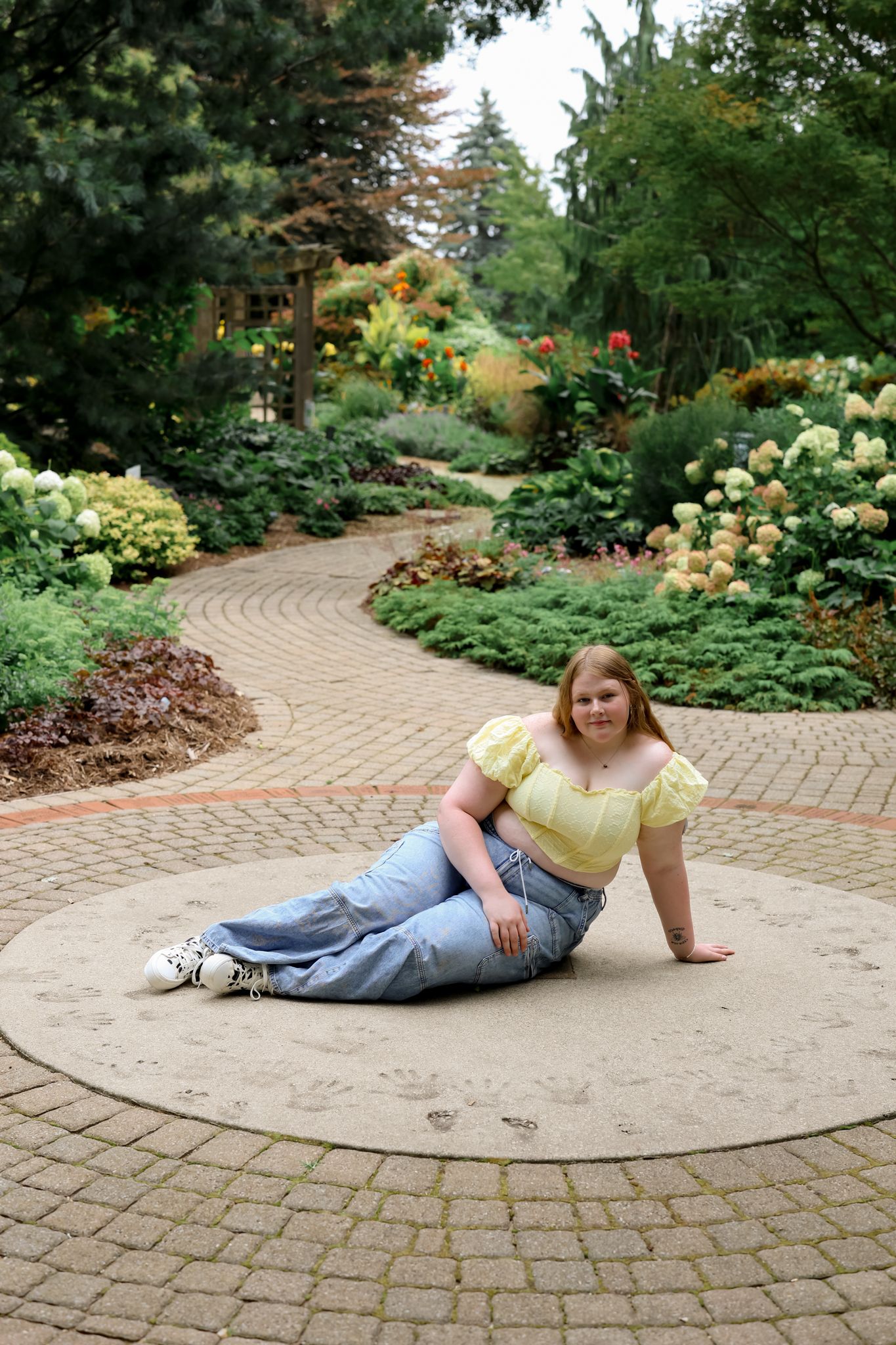 Person in yellow top and jeans posing on circular brick patio surrounded by garden.