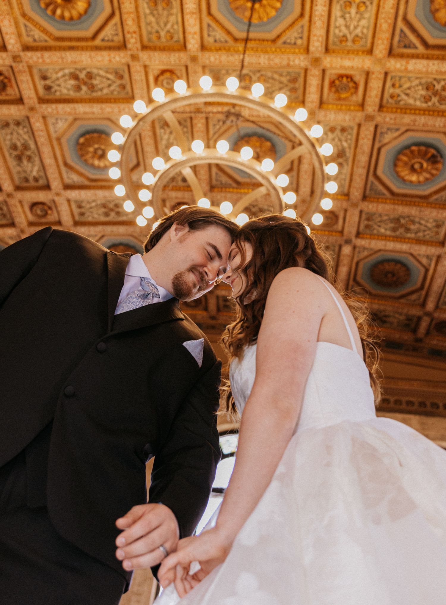 A bride and groom hold hands beneath an ornate gilded ceiling with a glowing chandelier in an elegant wedding venue.