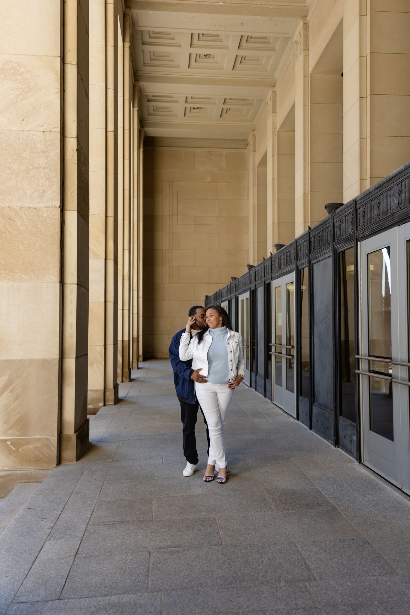 Couple walking through elegant stone colonnade corridor with tall columns and decorative ceiling.