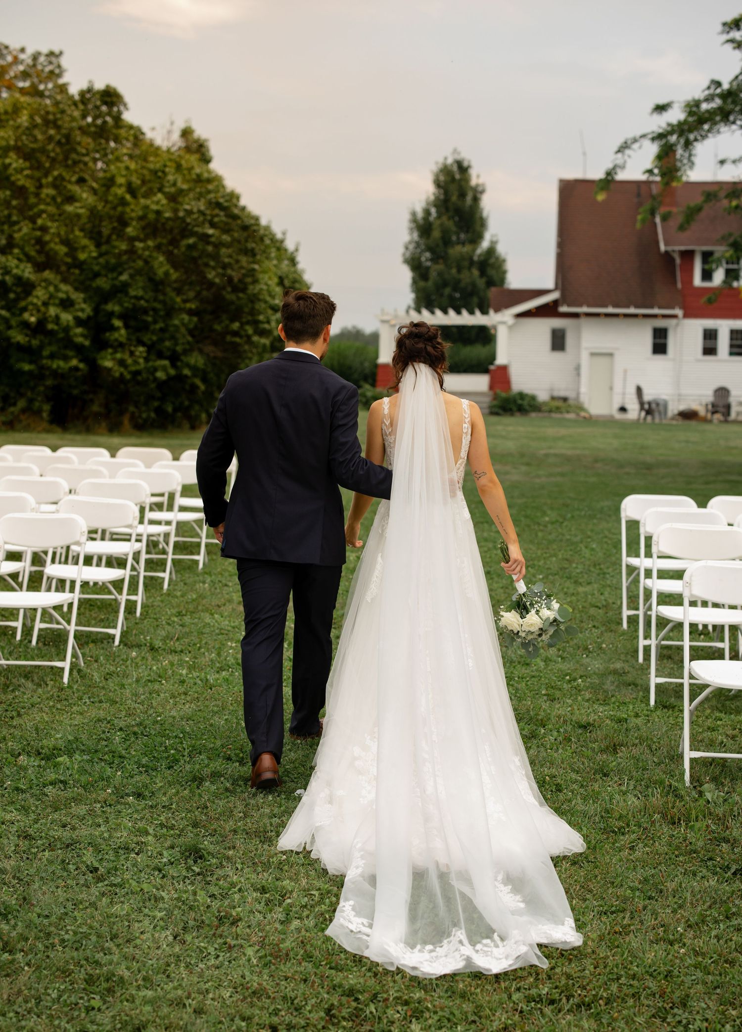 Couple walking through empty white chairs set up for outdoor wedding ceremony.