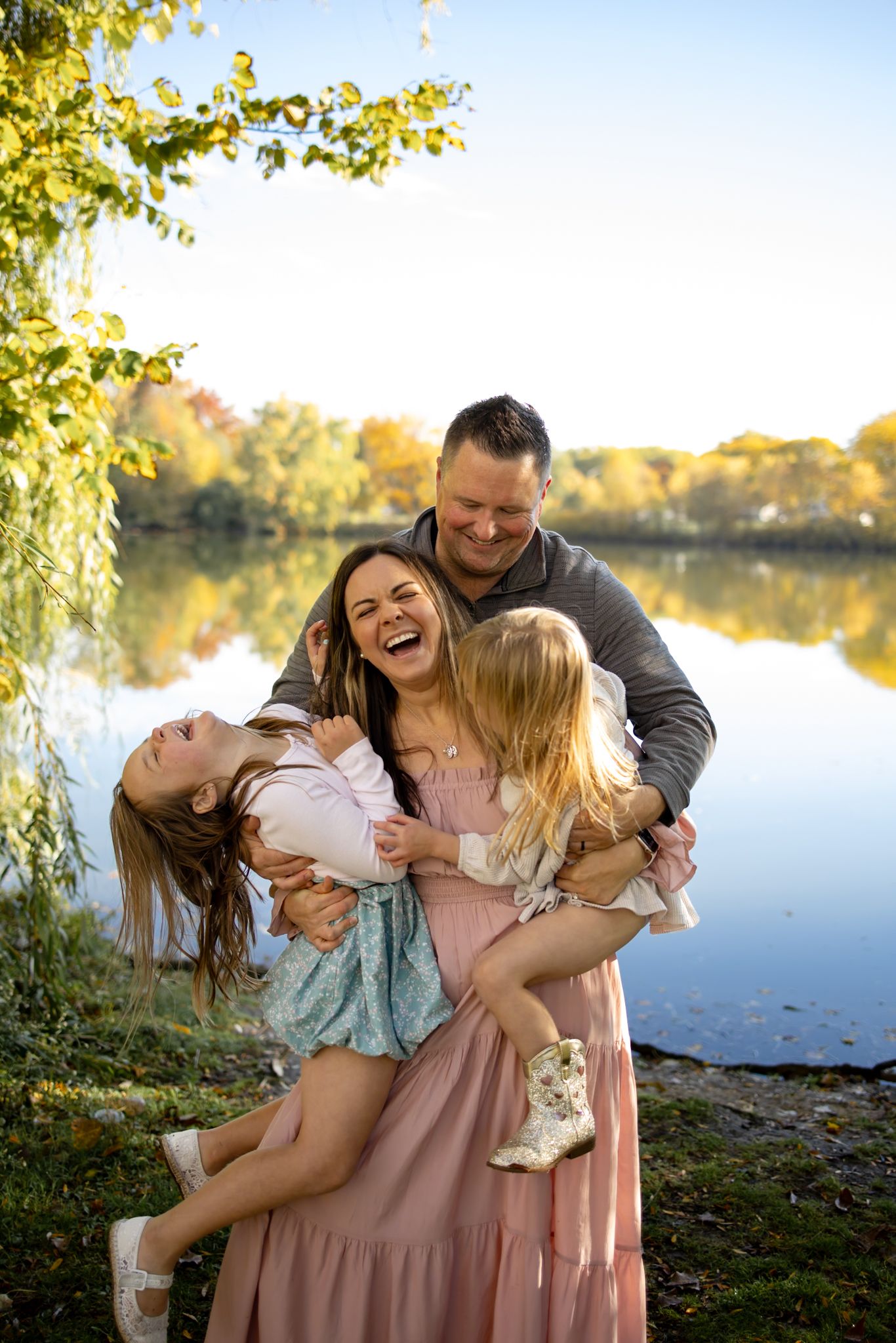 Family plays and laughs together during outdoor photo session by the lake.