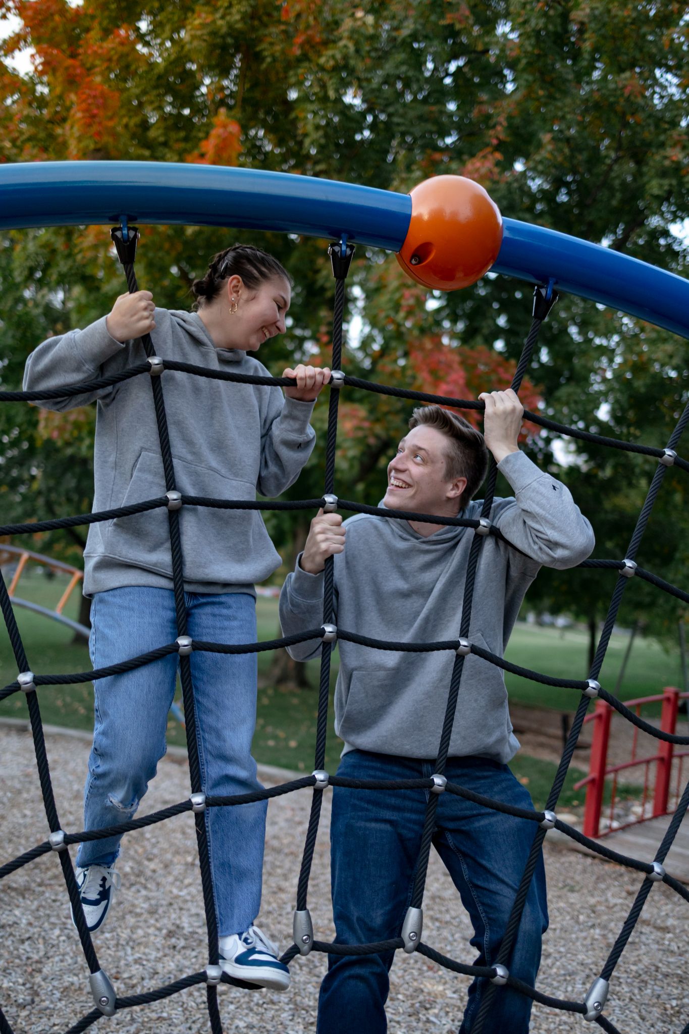 Two people in matching gray hoodies playfully interact on playground equipment during autumn.