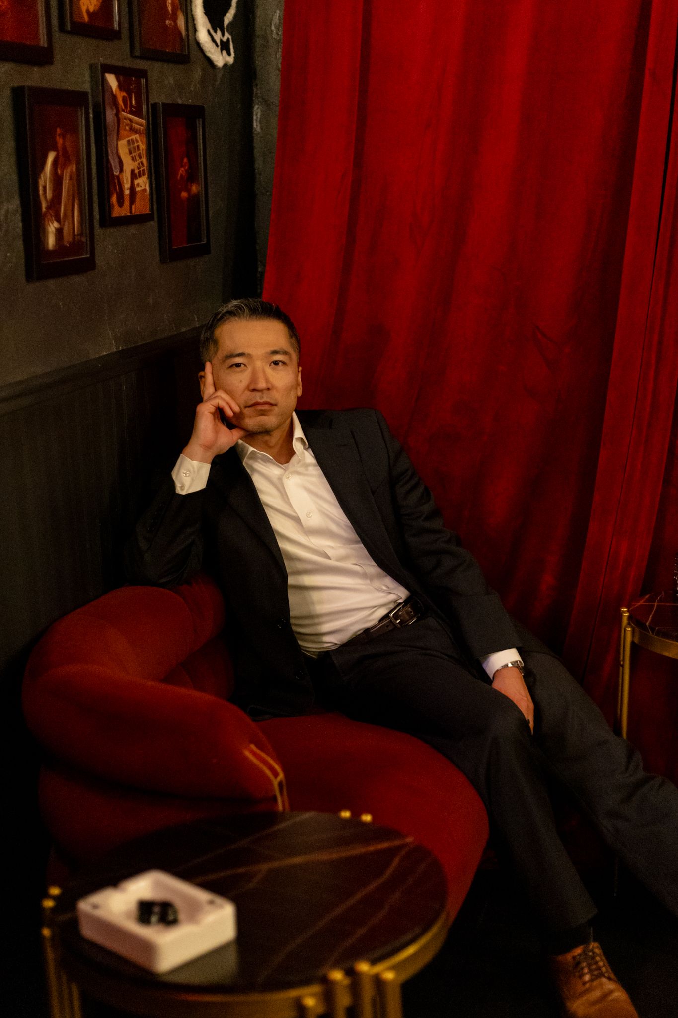 A person in formal attire reclines on a red velvet couch in a dimly lit room with framed artwork on the walls.
