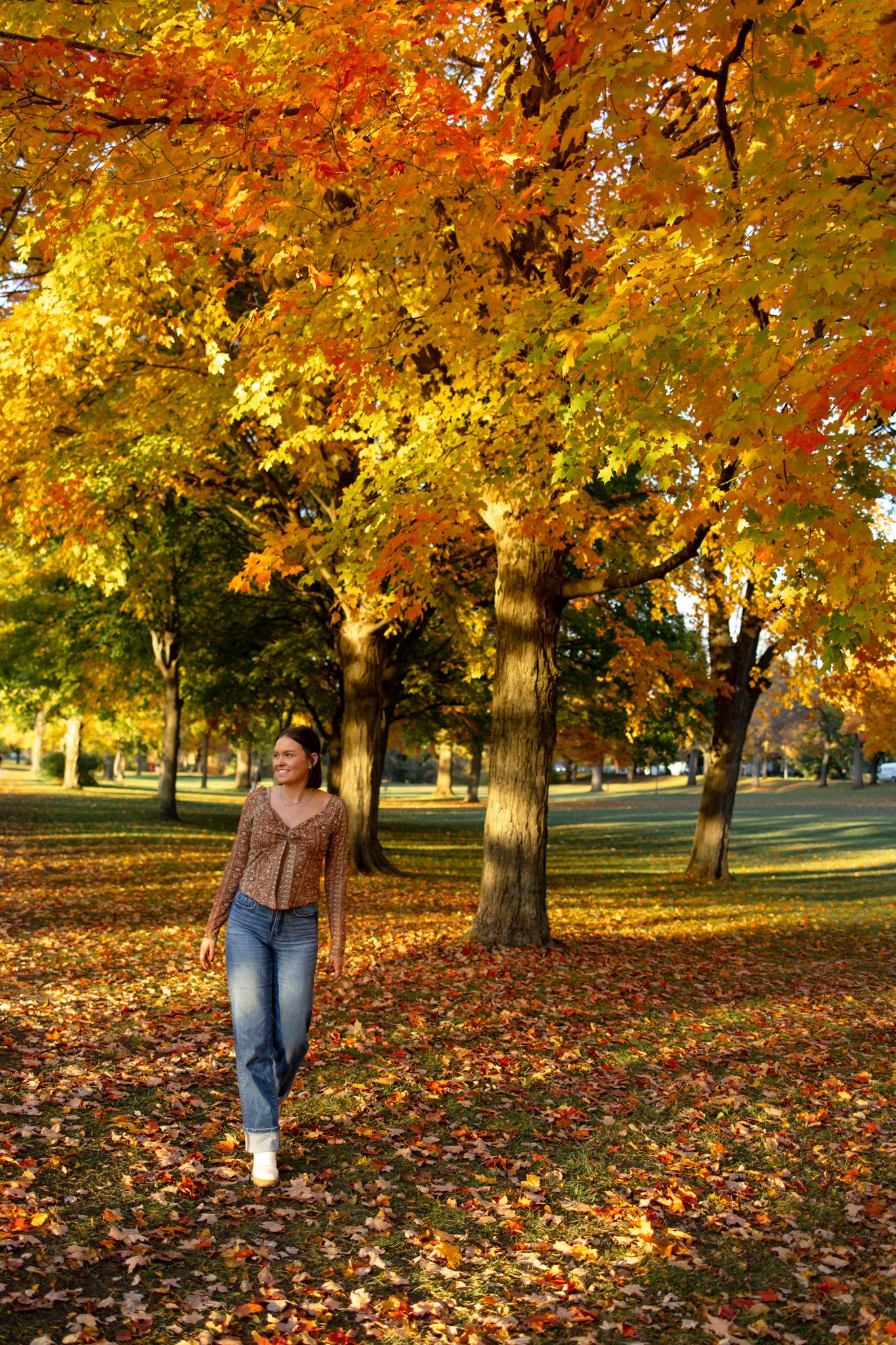 Person walking through a park with vibrant orange and yellow autumn leaves scattered on the ground.