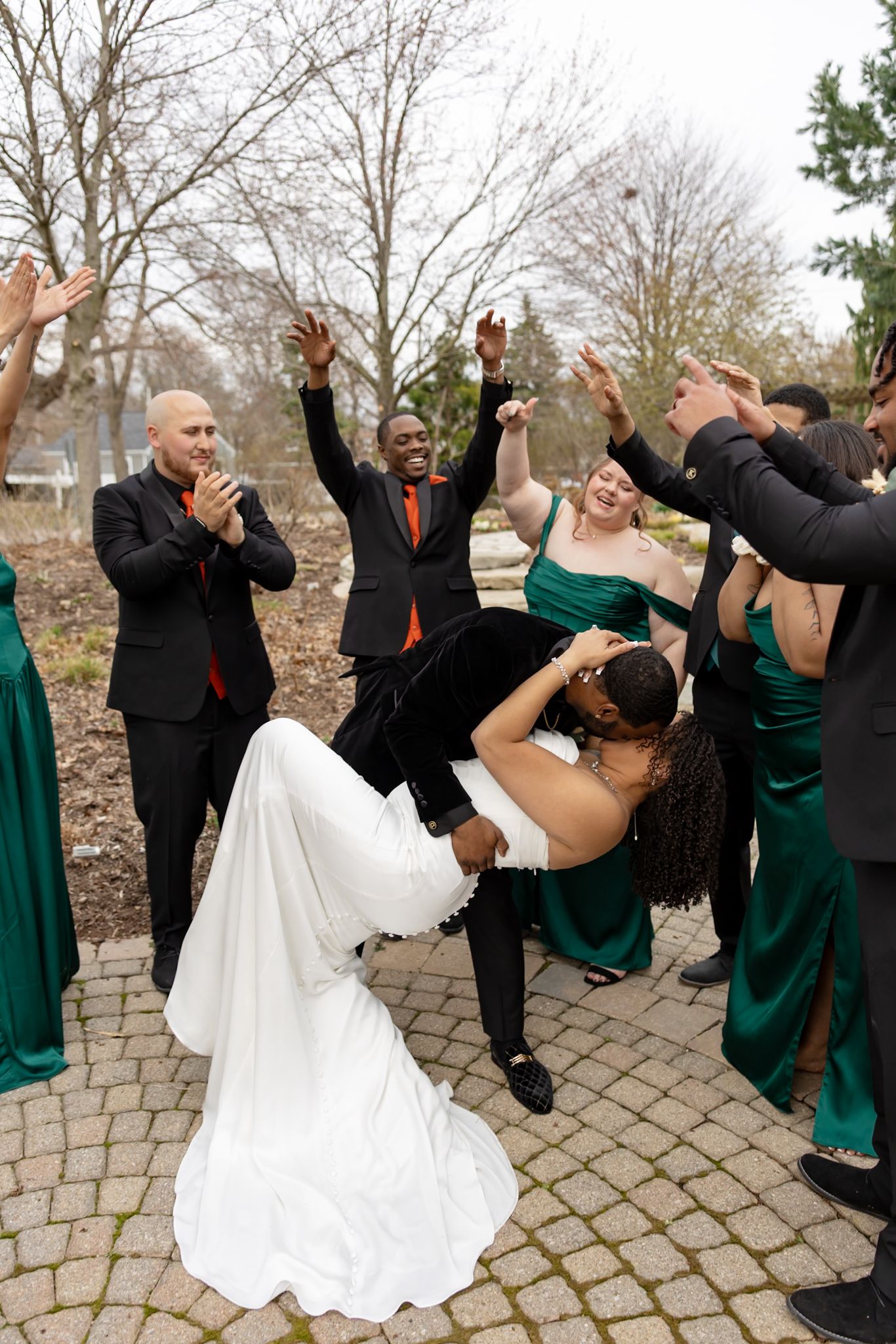 Bride and groom share a romantic dip kiss surrounded by cheering wedding party in green dresses and black suits outdoors.