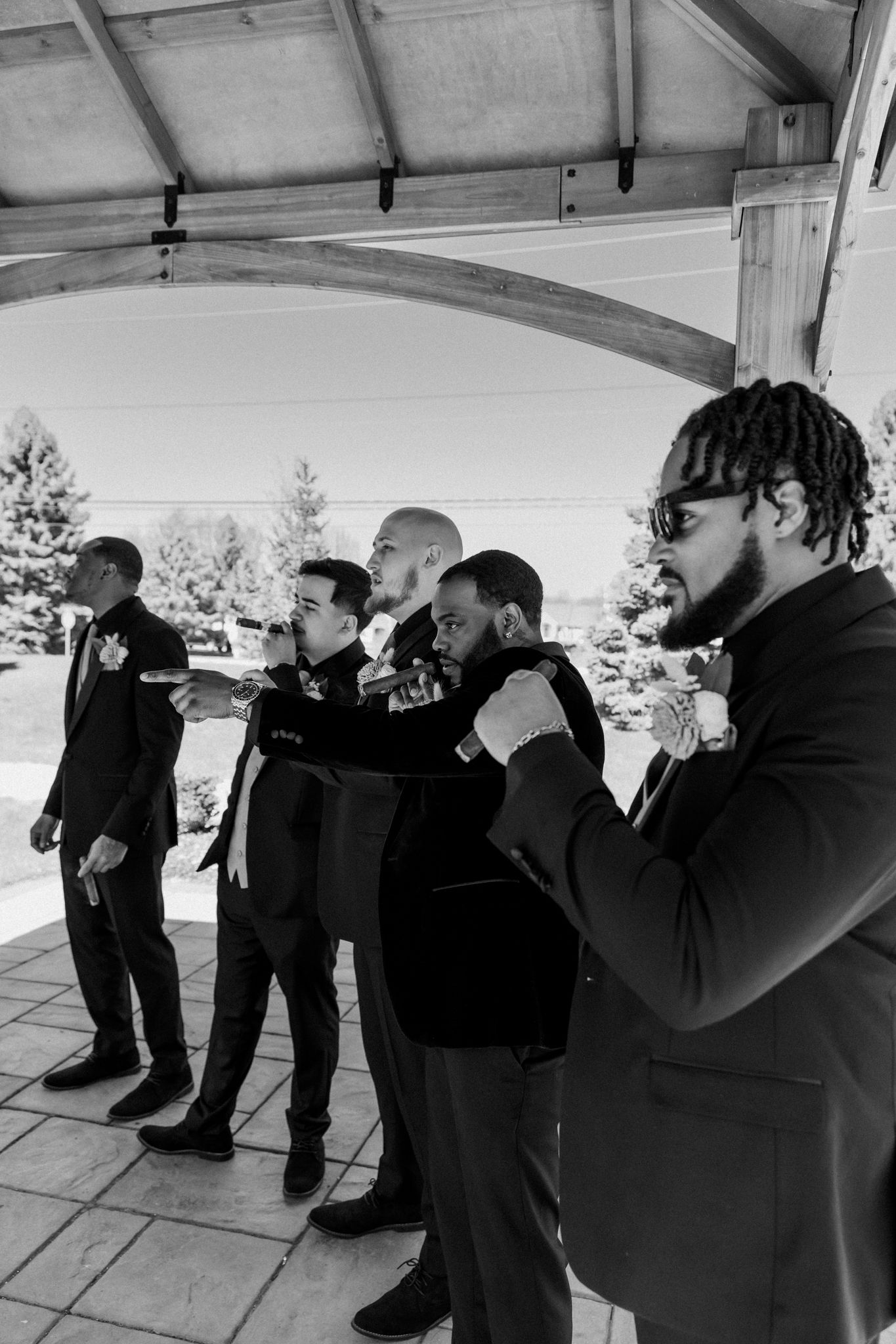 Black and white photo of groomsmen in suits adjusting ties under a pavilion before a wedding ceremony.