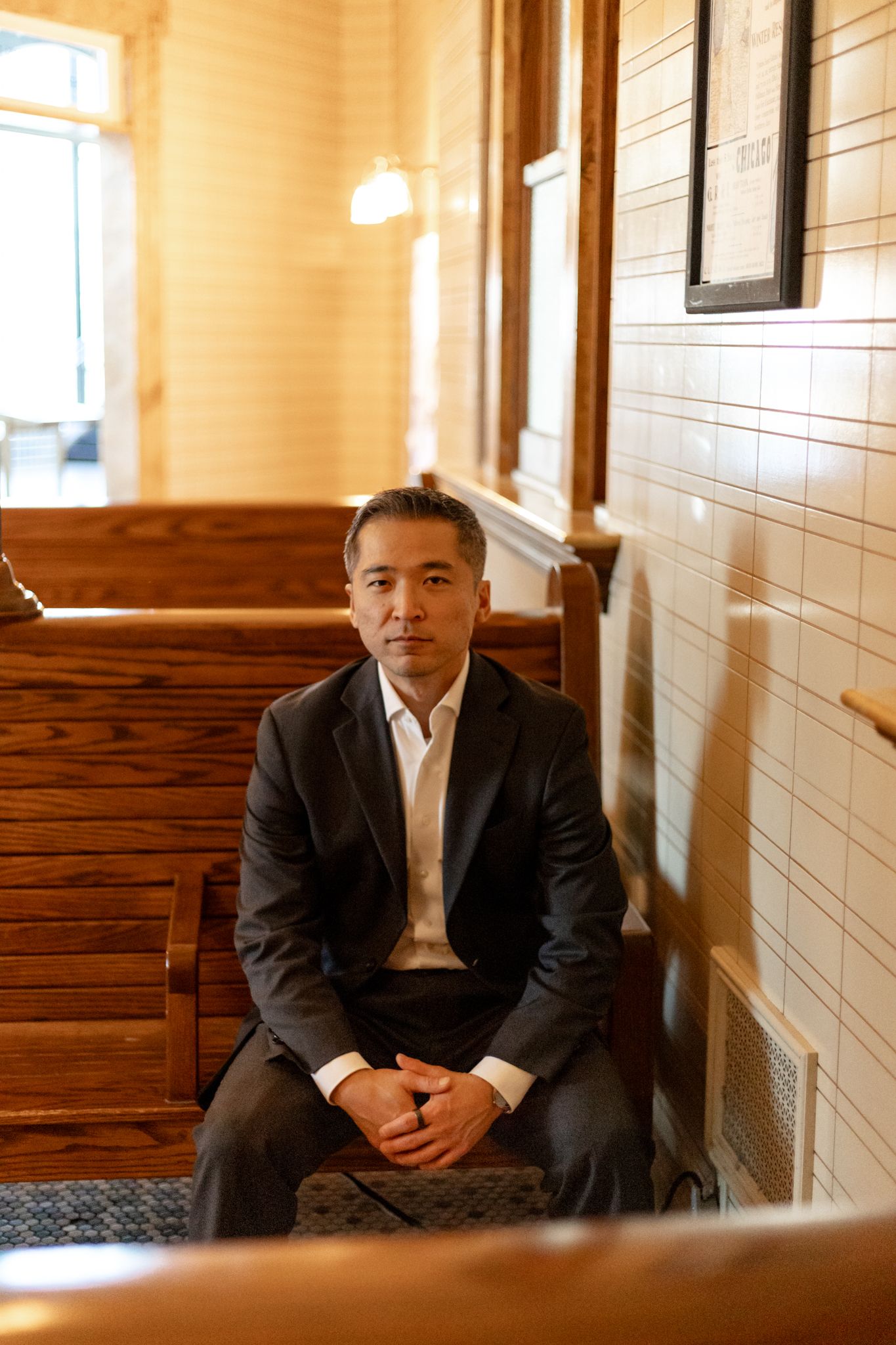 A person in a dark suit sits in a warmly lit hallway with white tiled walls and wooden benches.