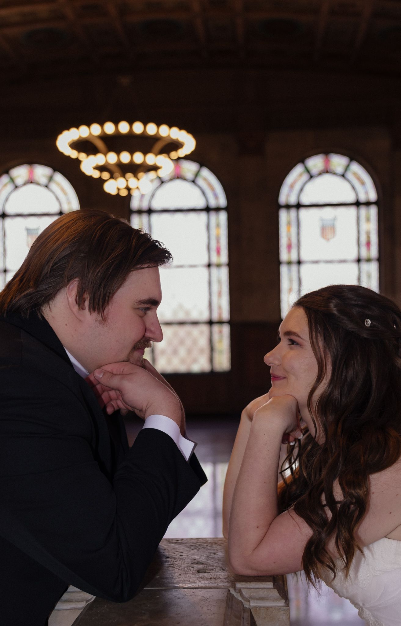 Newlywed couple gazing lovingly at each other at table with elegant stained glass windows and chandelier backdrop.