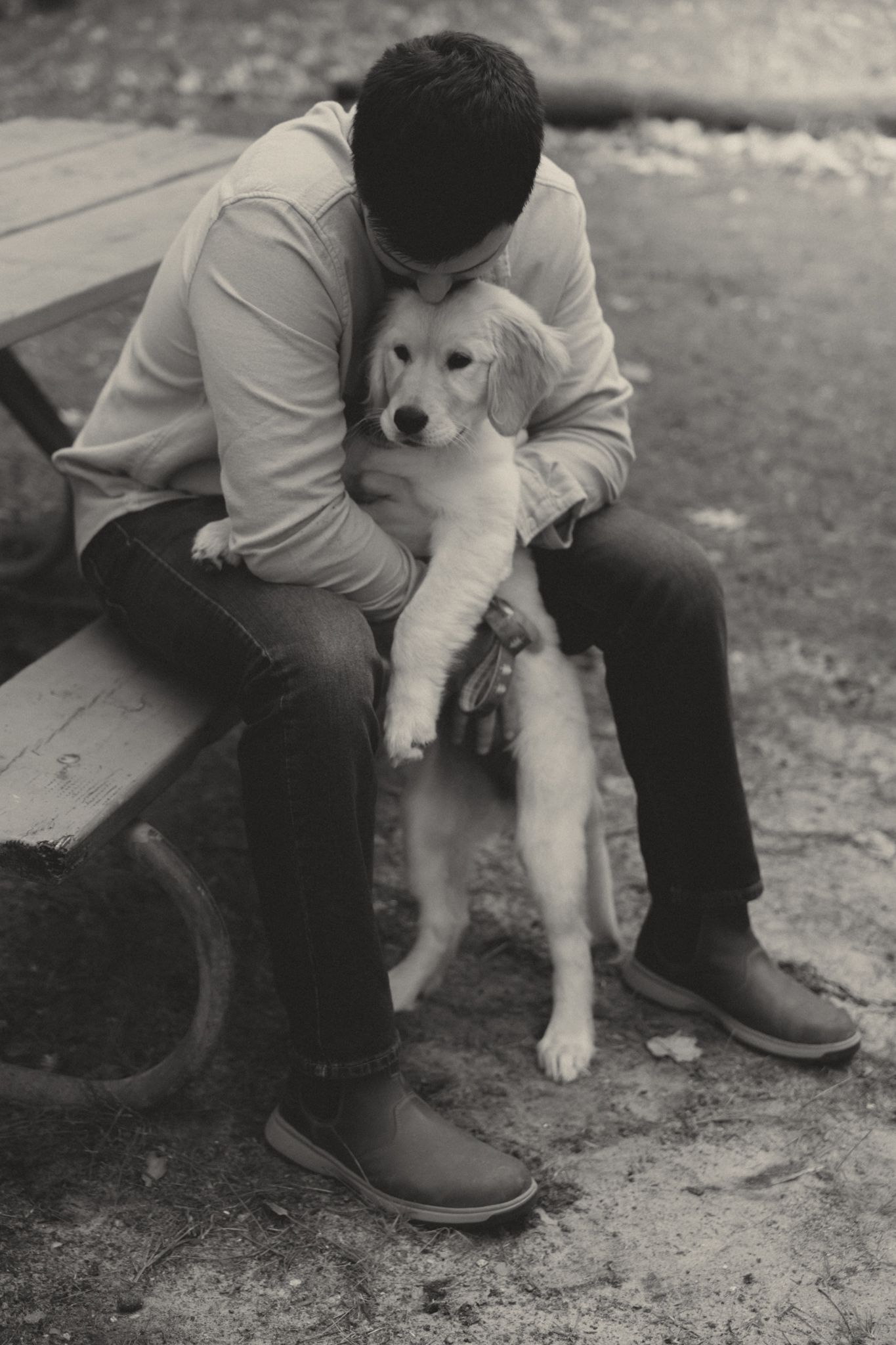 Black and white photo of a young man sitting on a bench, tenderly hugging a golden retriever puppy outdoors.
