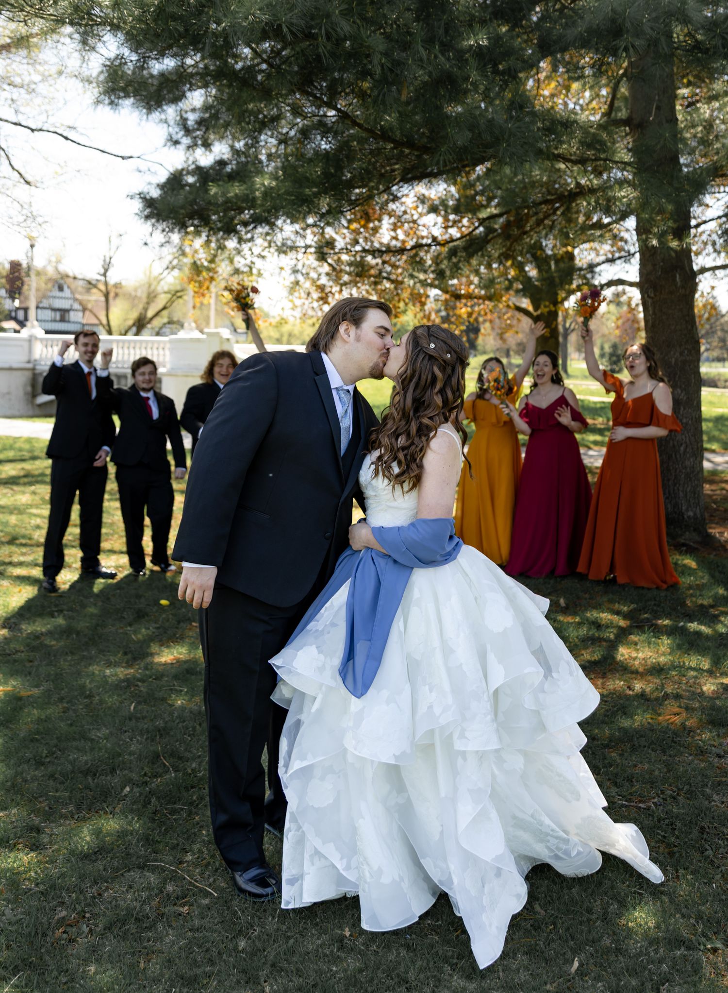 Bride and groom share a kiss outdoors while wedding party in colorful dresses watches during fall ceremony.