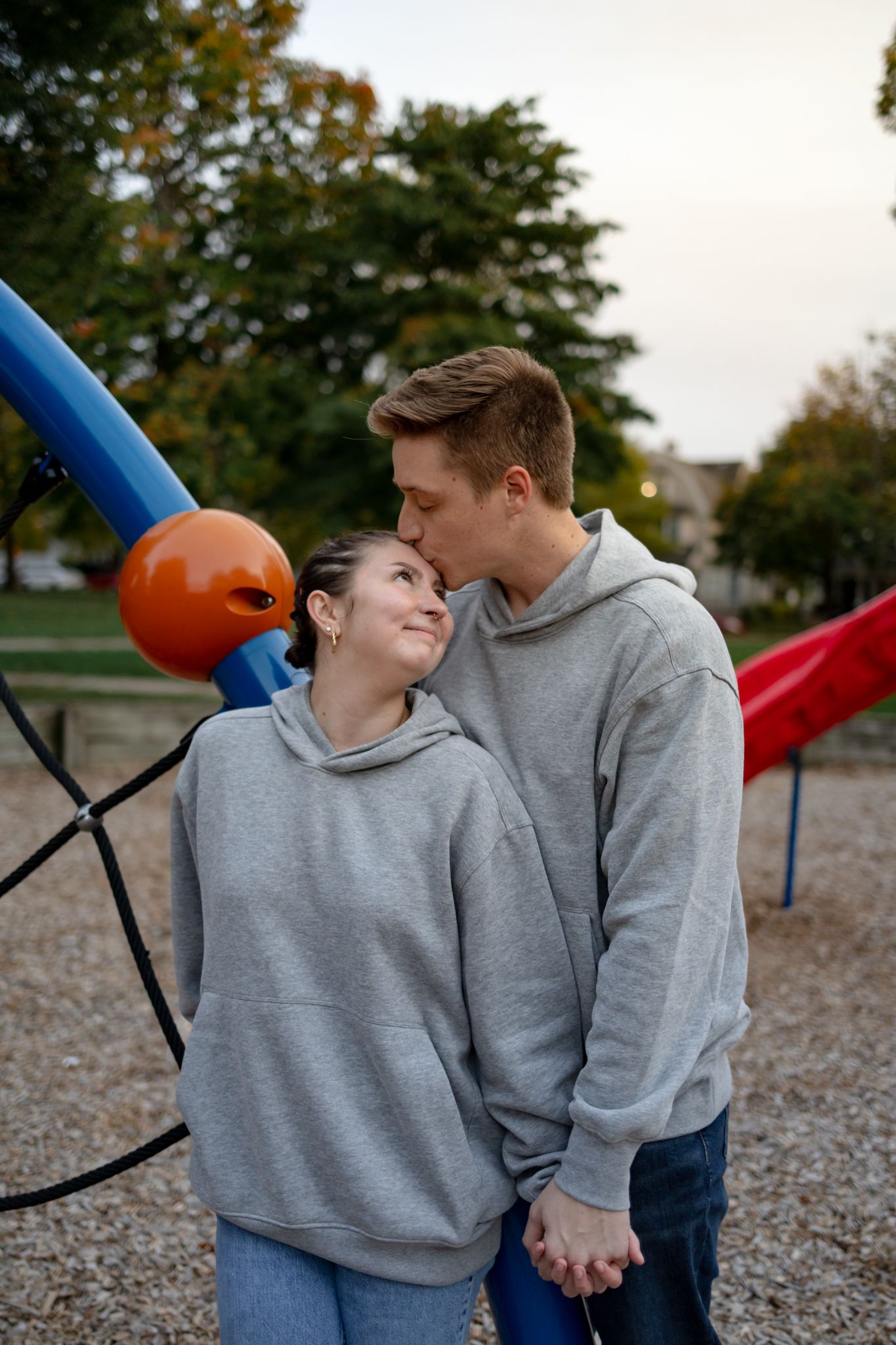 Two people wearing matching gray hoodies share a tender moment at a playground.