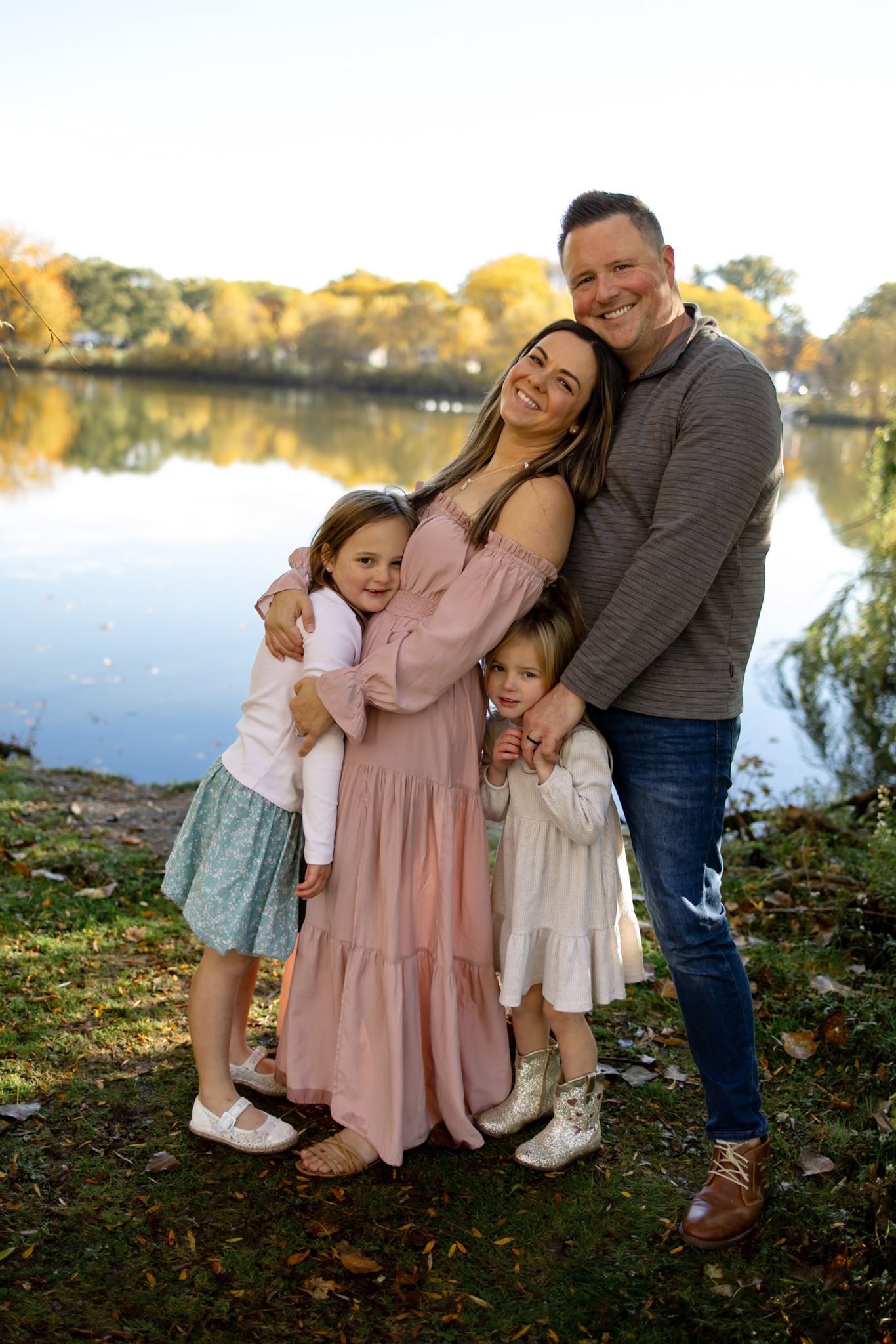 A family shares joyful moments together by a lake during an autumn photoshoot in coordinated pink and grey outfits.