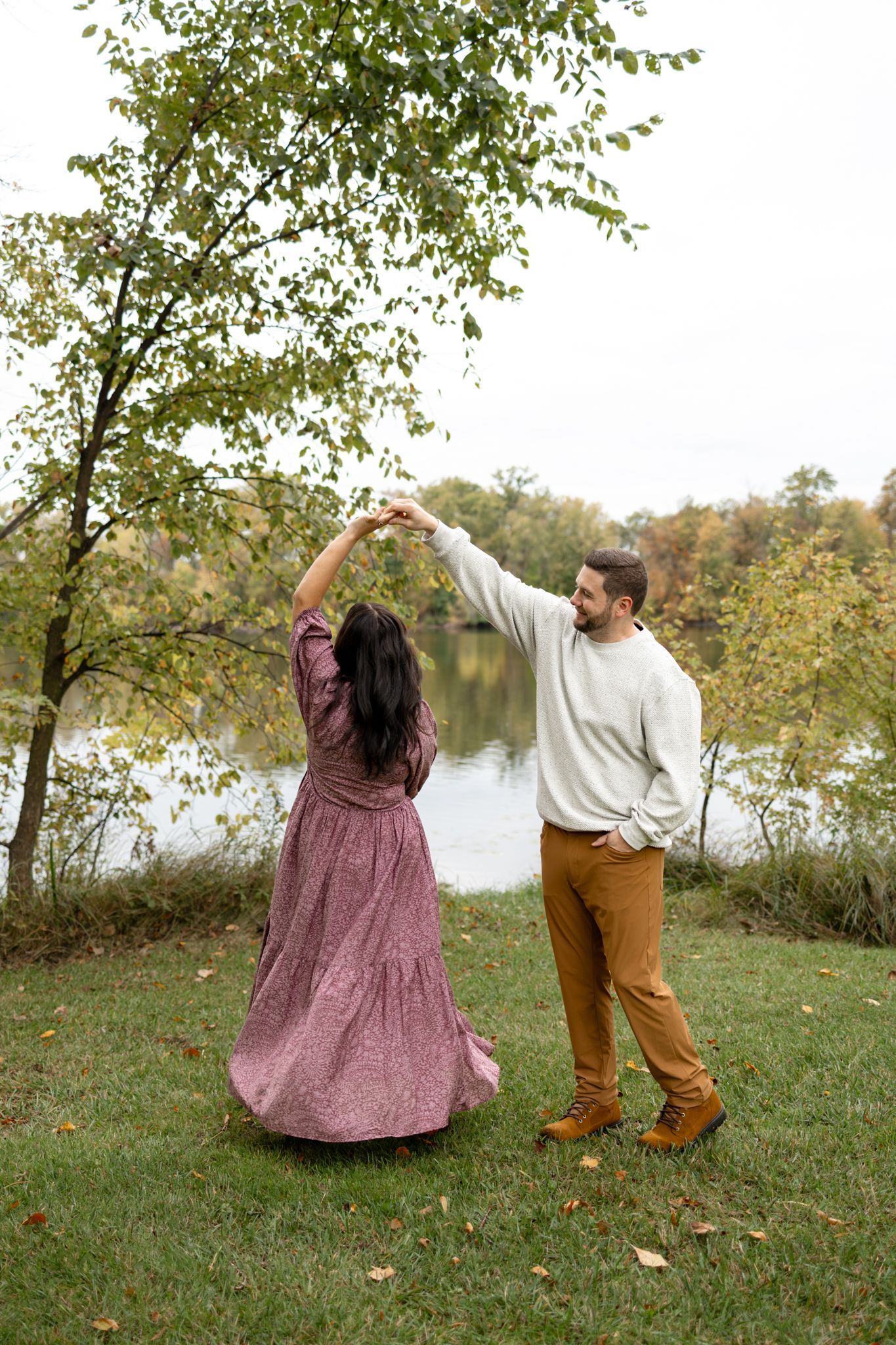 A couple dances playfully on grass near a lake during autumn with one wearing a flowy mauve dress.