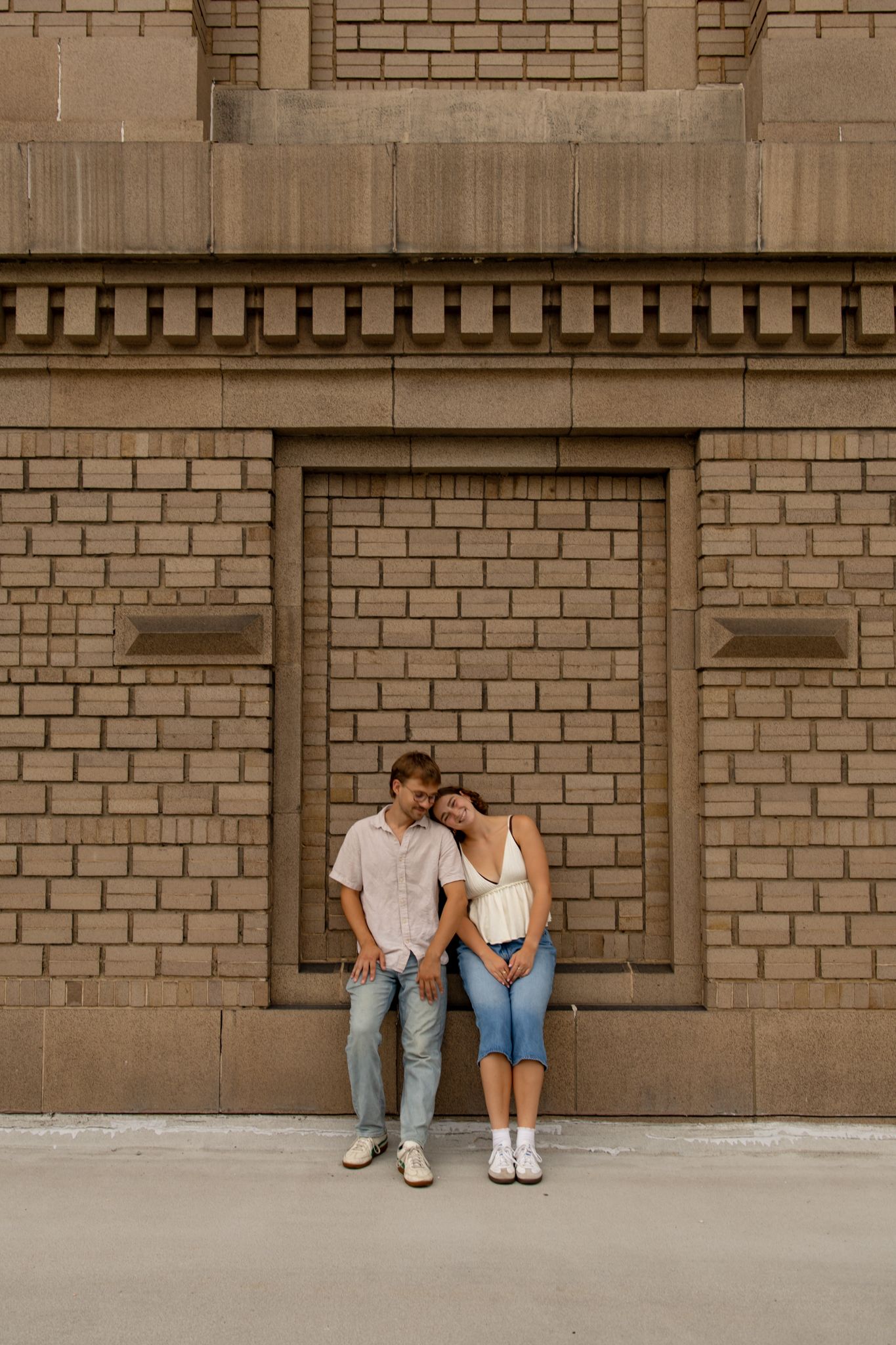 Couple sitting together against brick wall in casual summer outfits.