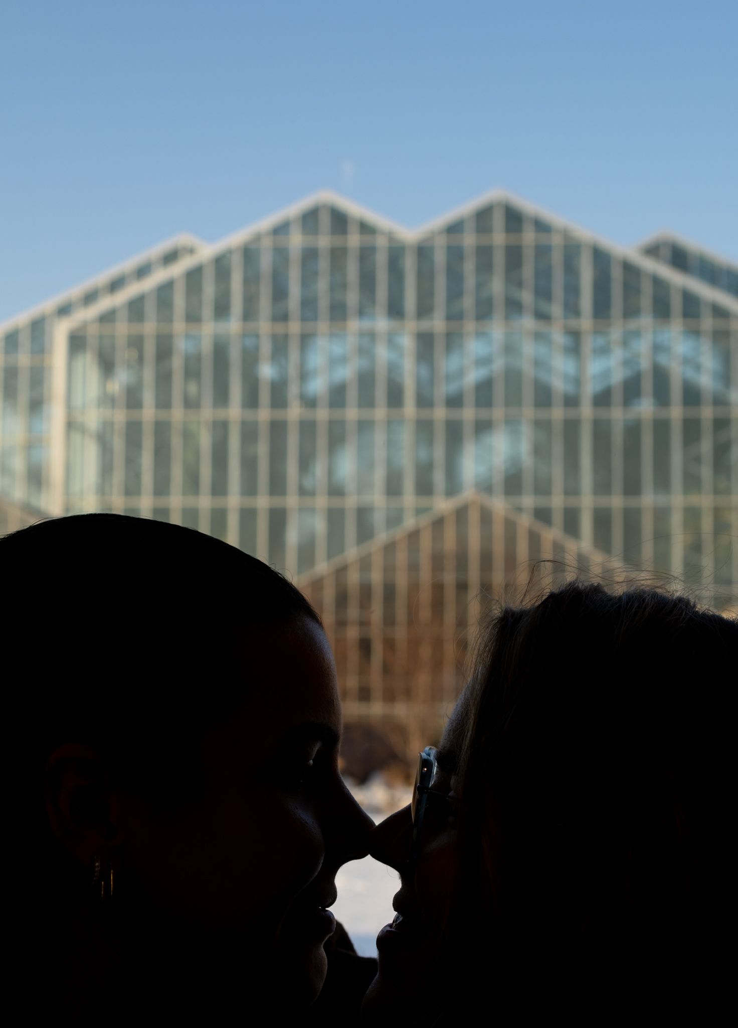 Silhouettes of two figures stand in front of large greenhouse windows during sunset, creating a dramatic backlit effect.
