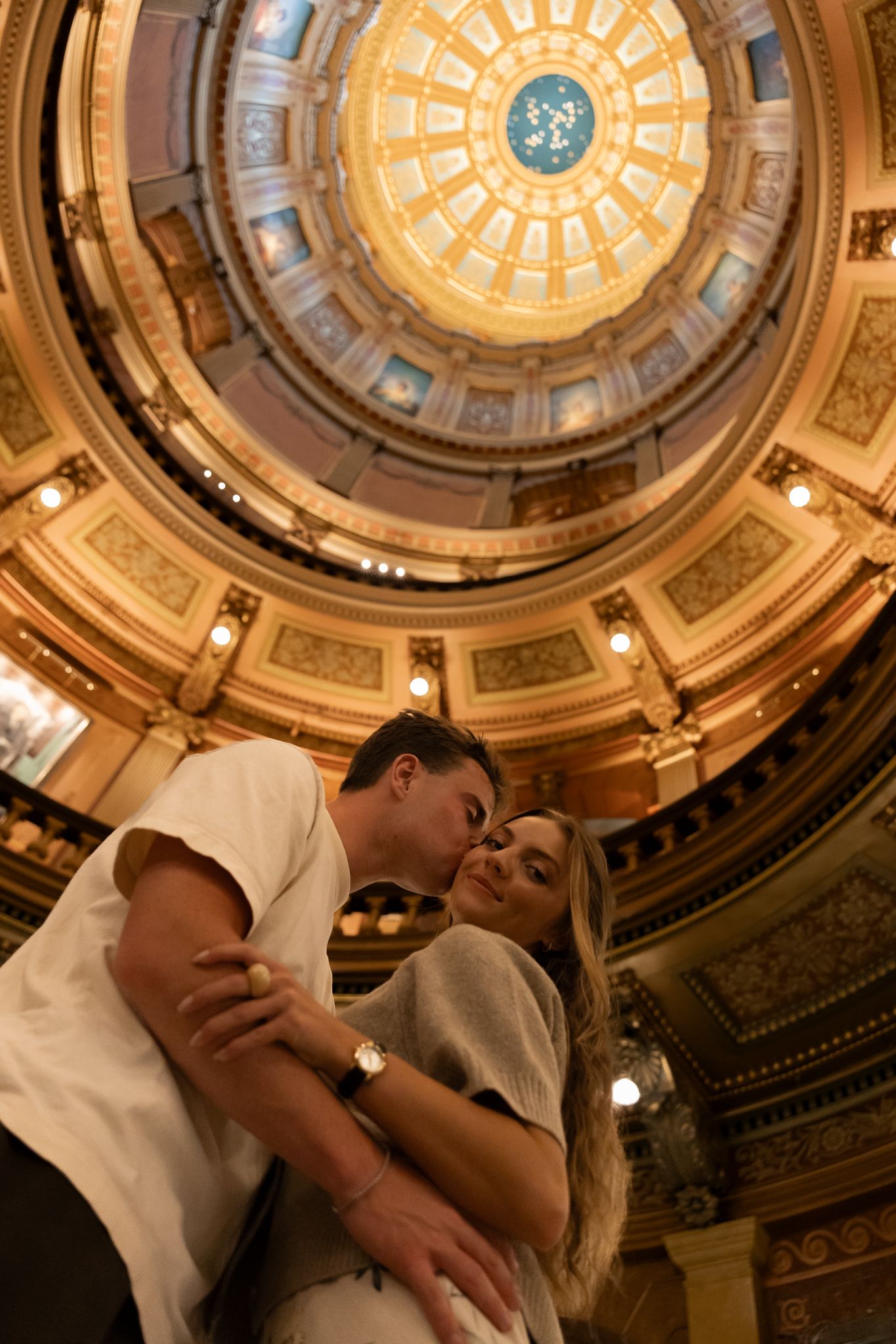 Romantic couple sharing a kiss under an ornate golden domed ceiling in a grand historic building.