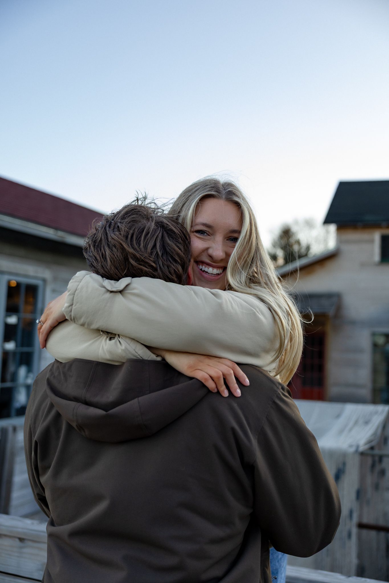 A smiling blonde woman hugging a person in a dark jacket outdoors near rustic buildings at dusk.