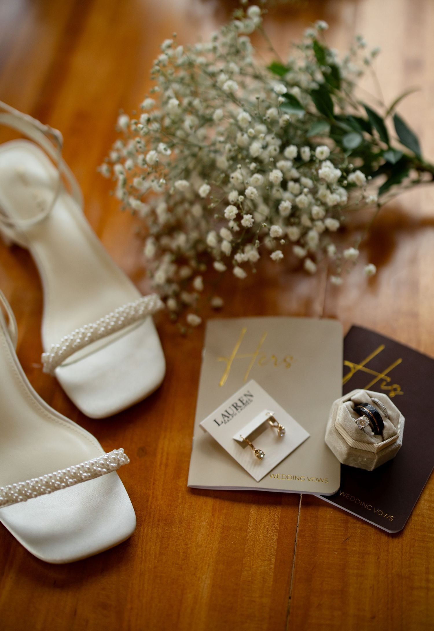Wedding details with white shoes, baby's breath flowers, and rings on wooden surface.
