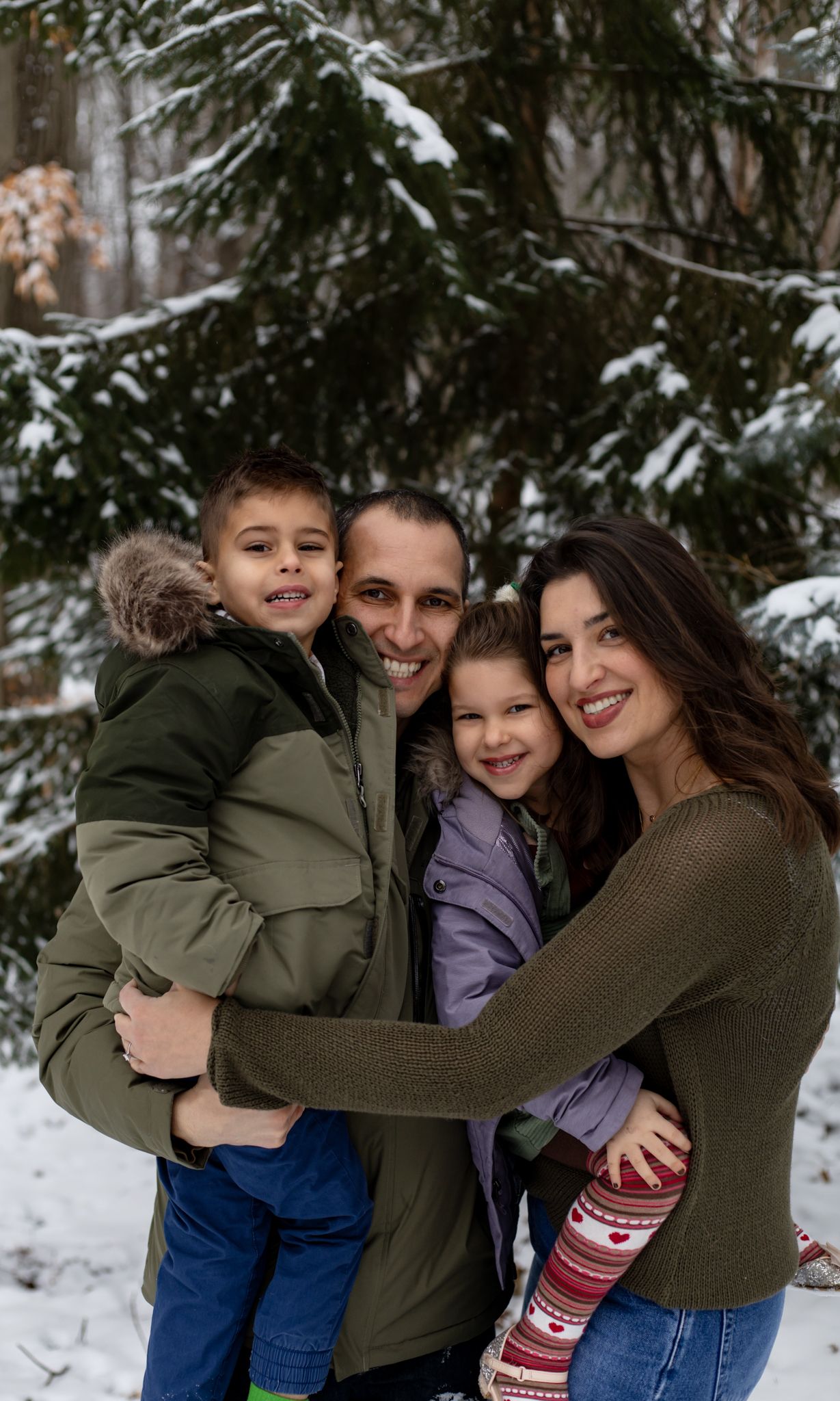 Winter family photoshoot with snow-covered evergreen trees in background.