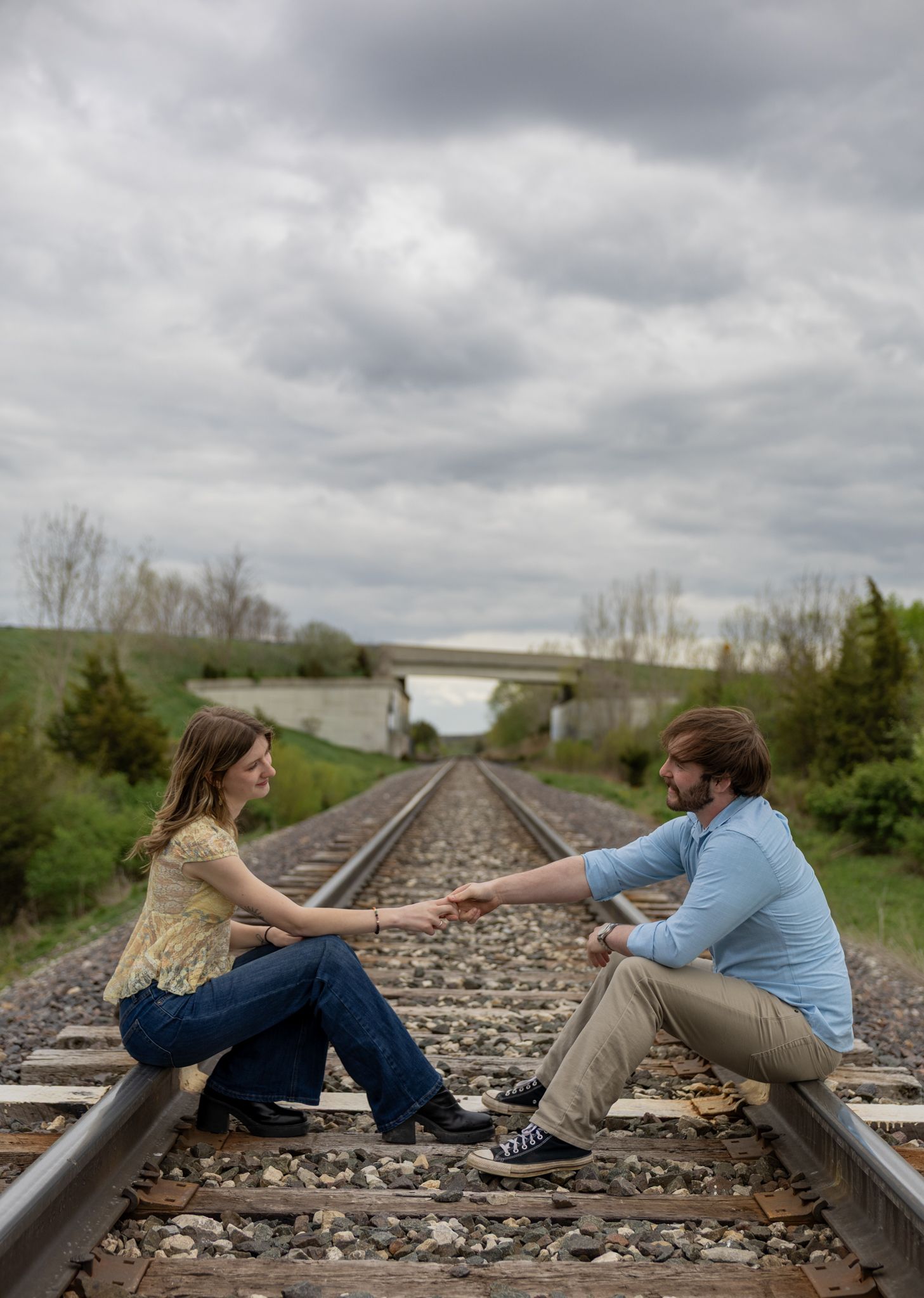 Couple sitting face-to-face on railroad tracks reaching toward each other under cloudy skies.