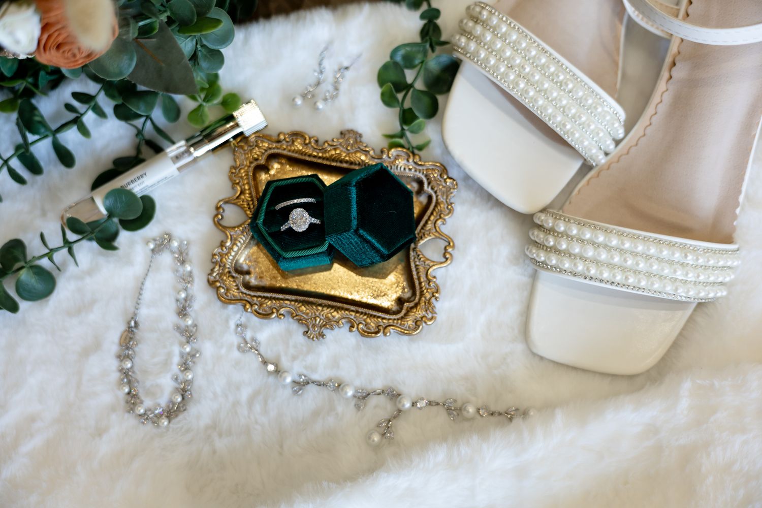 Bridal flat lay with diamond ring in green velvet box, pearl heels, crystal bracelet, and gold tray on white fur.
