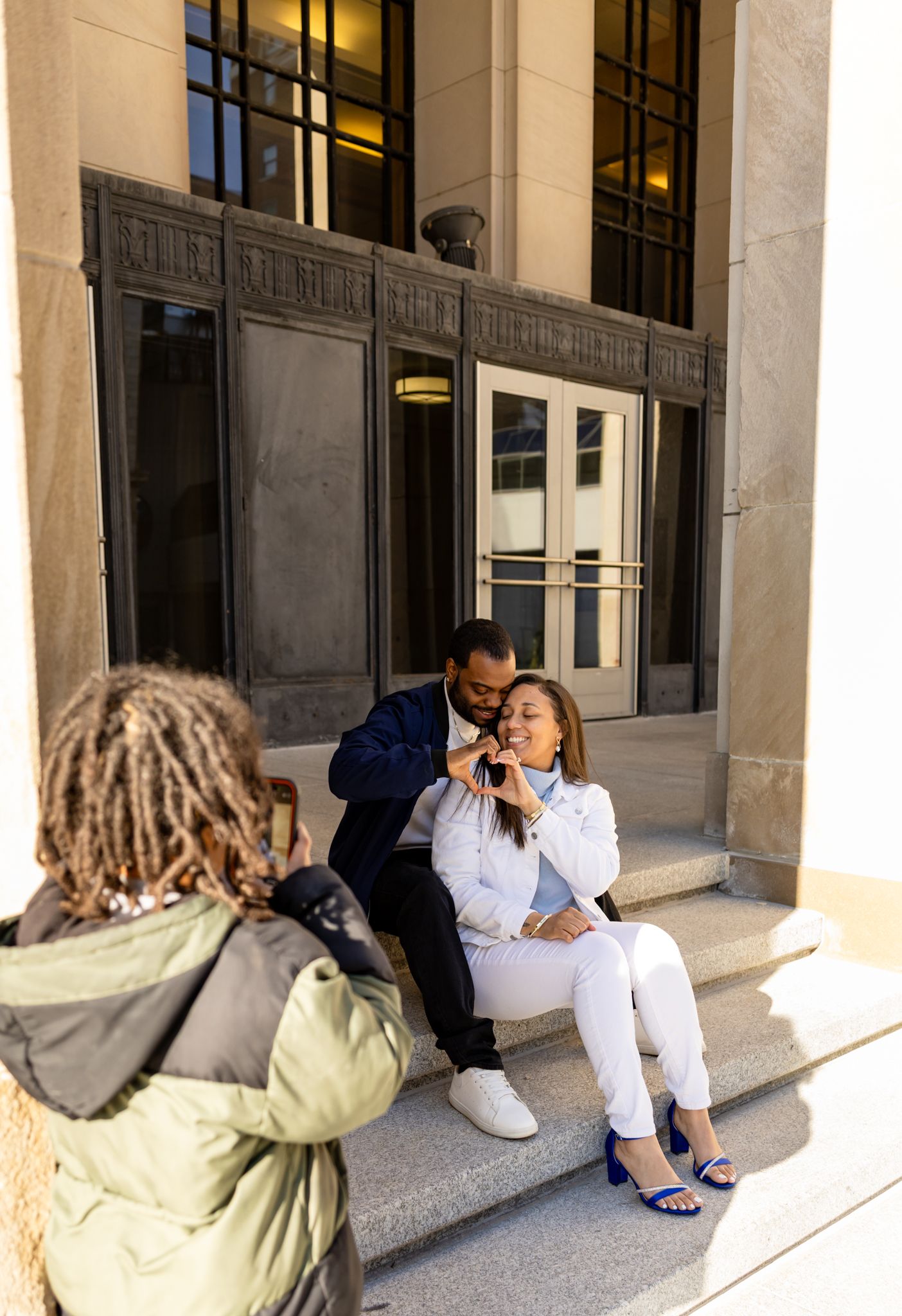 A couple poses on building steps while a photographer captures their romantic moment outdoors.