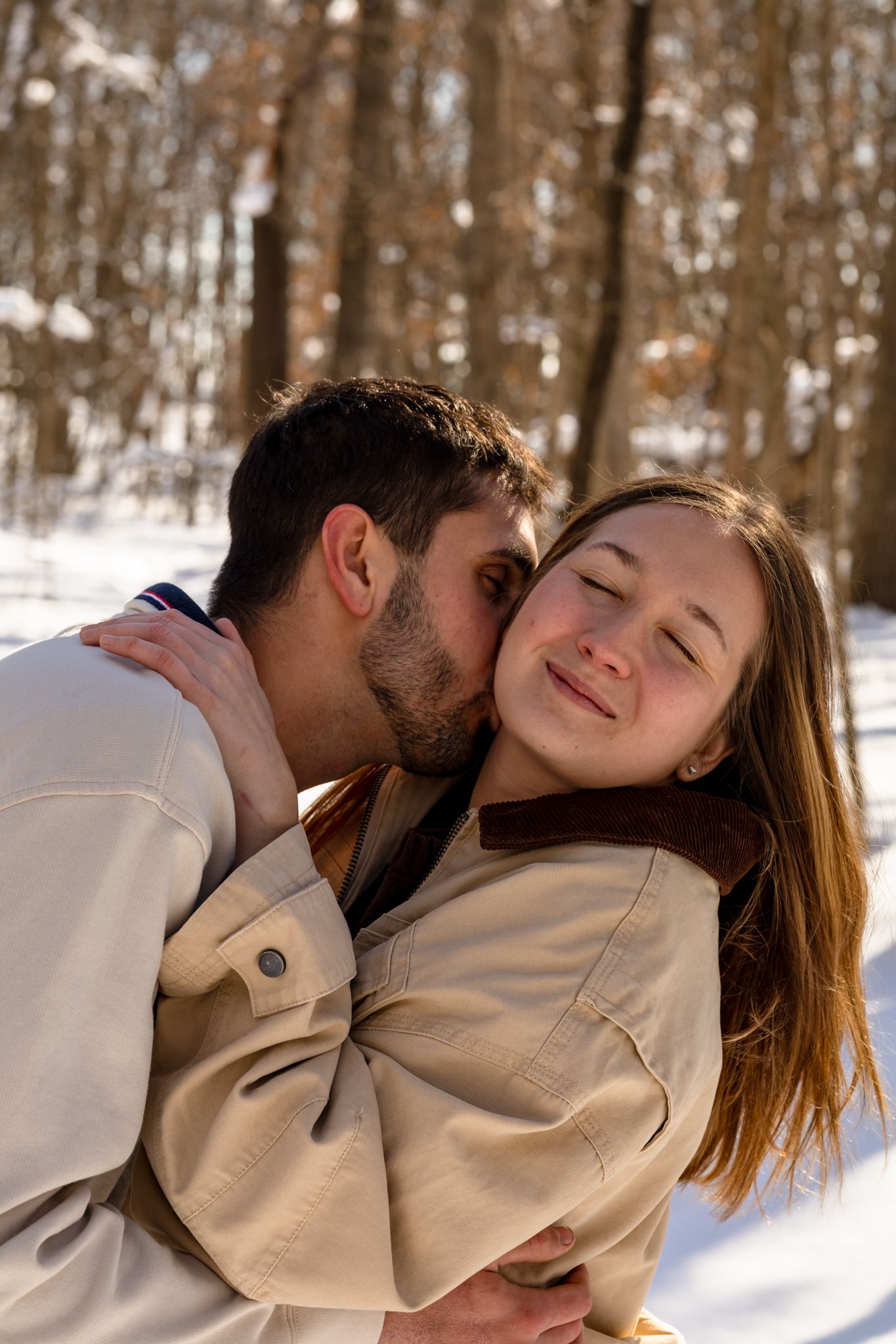 A couple embraces in a snowy winter forest, man kissing woman on cheek as she smiles with eyes closed.