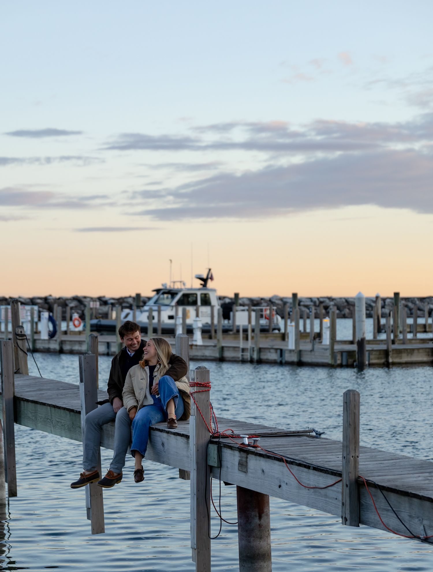 Couple sitting together on a wooden dock at sunset with boats moored in a marina behind them.