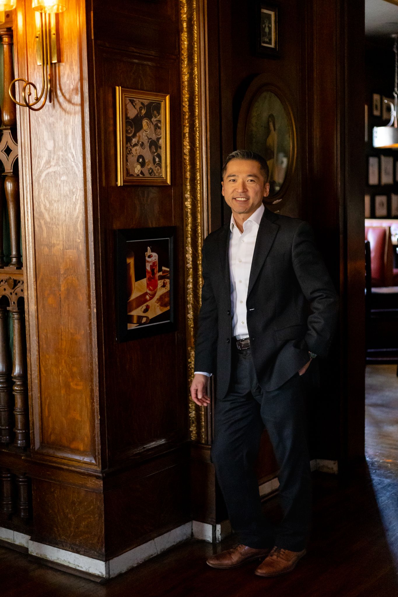 A man in a dark suit stands in an elegant wood-paneled hallway with ornate gold trim and decorative details.